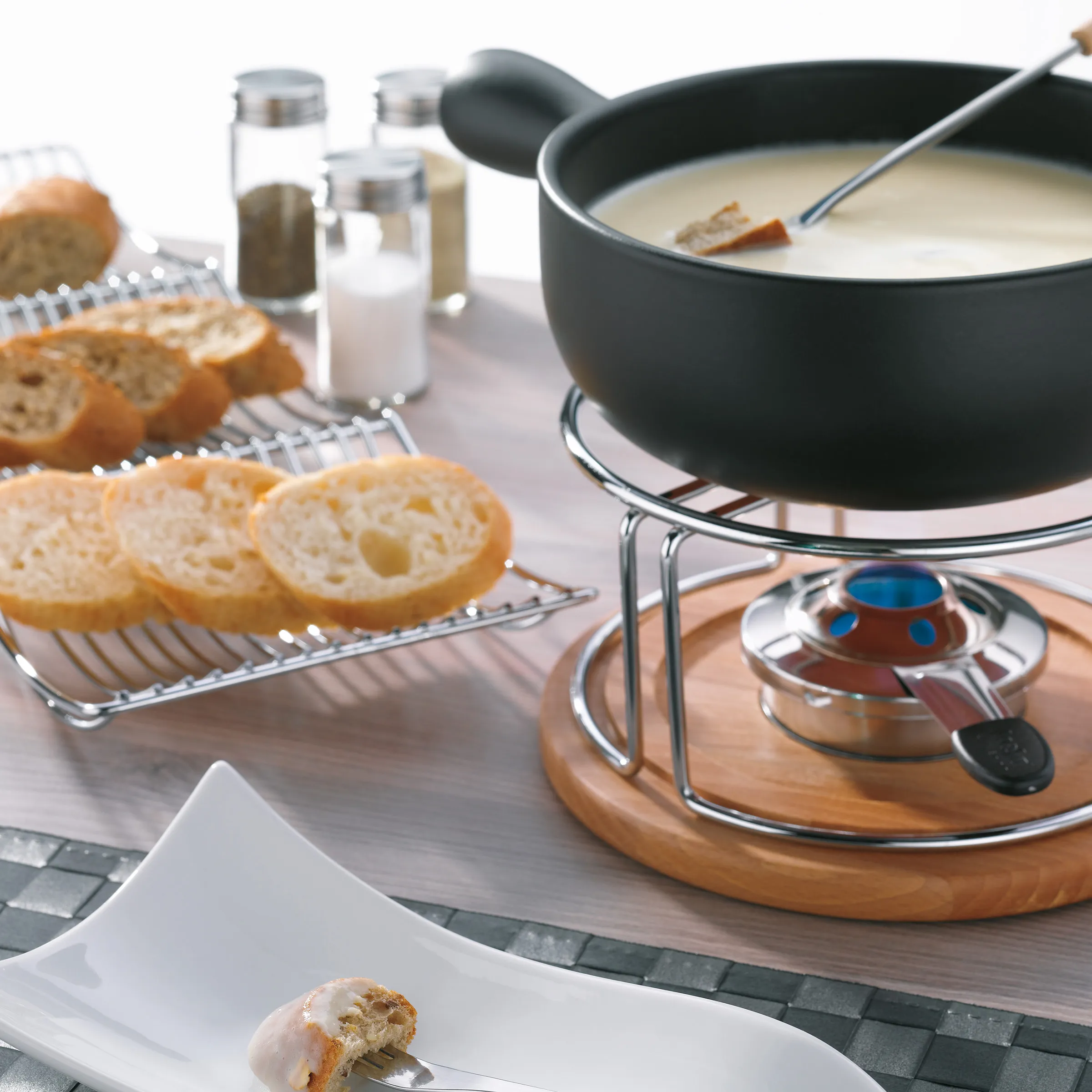 Fondue Set - Käsefondue Zubehör kaufen Fondue-Set mit schwarzem Topf auf Holzständer, Brokkoli-Scheiben auf Tablett und Würzflaschen auf weißer Oberfläche