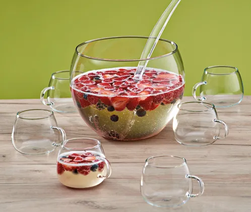 Beerengetränk Bowle - Fruchtsaft Servierset mit Gläsern Glasschüssel mit rotem Beerengetränk und Strohhalm, umgeben von leeren Trinkgläsern auf Holztisch vor grünem Hintergrund
