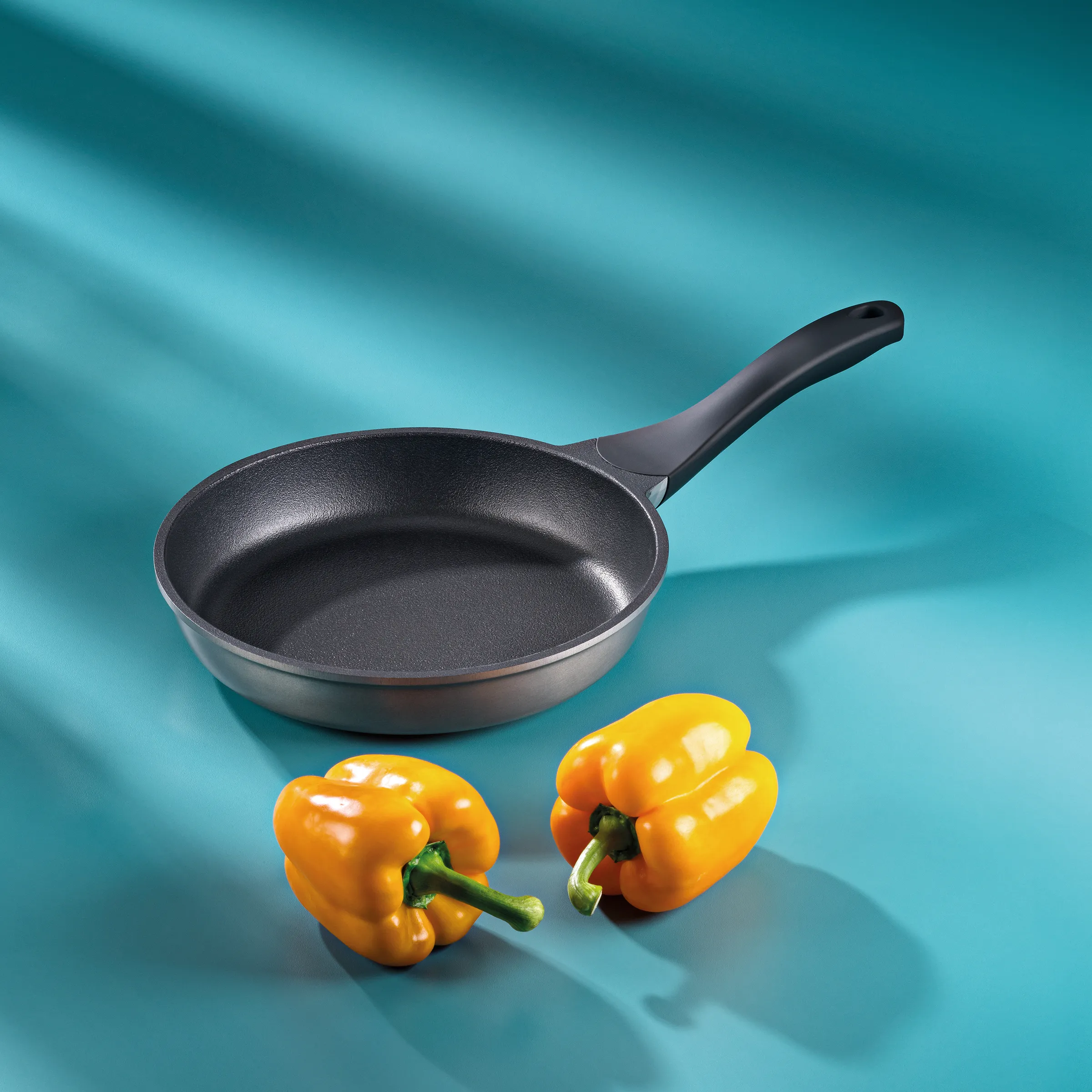Antihaft-Bratpfanne mit ergonomischem Griff - Hochwertige Küchenausstattung Graue Bratpfanne mit Antihaftbeschichtung und zwei gelben Paprika auf türkisem Hintergrund