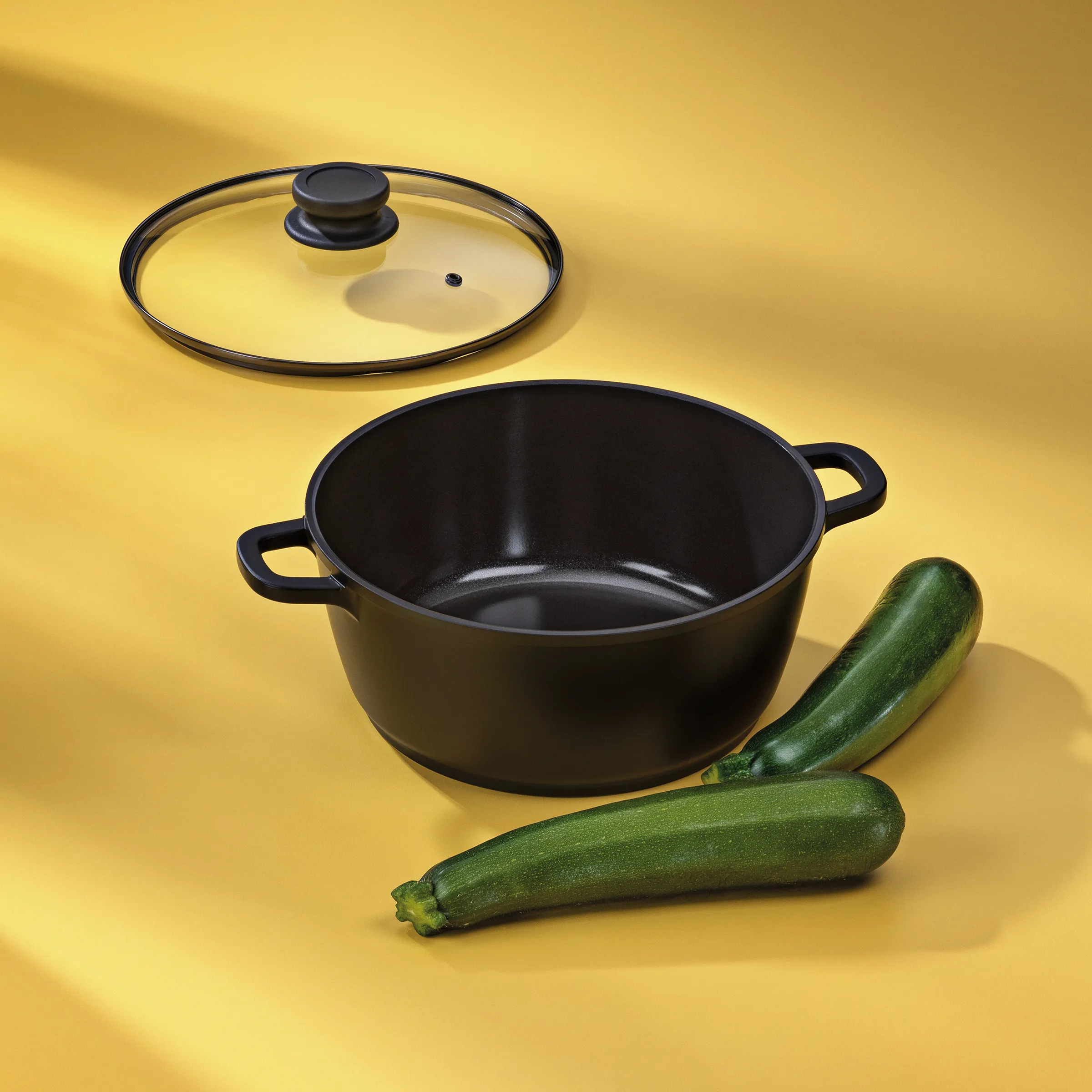 Kochtopf mit Deckel und Zucchini - Kochgeschirr Set Schwarzer Kochtopf mit Glasdeckel und zwei grüne Zucchini auf gelbem Hintergrund