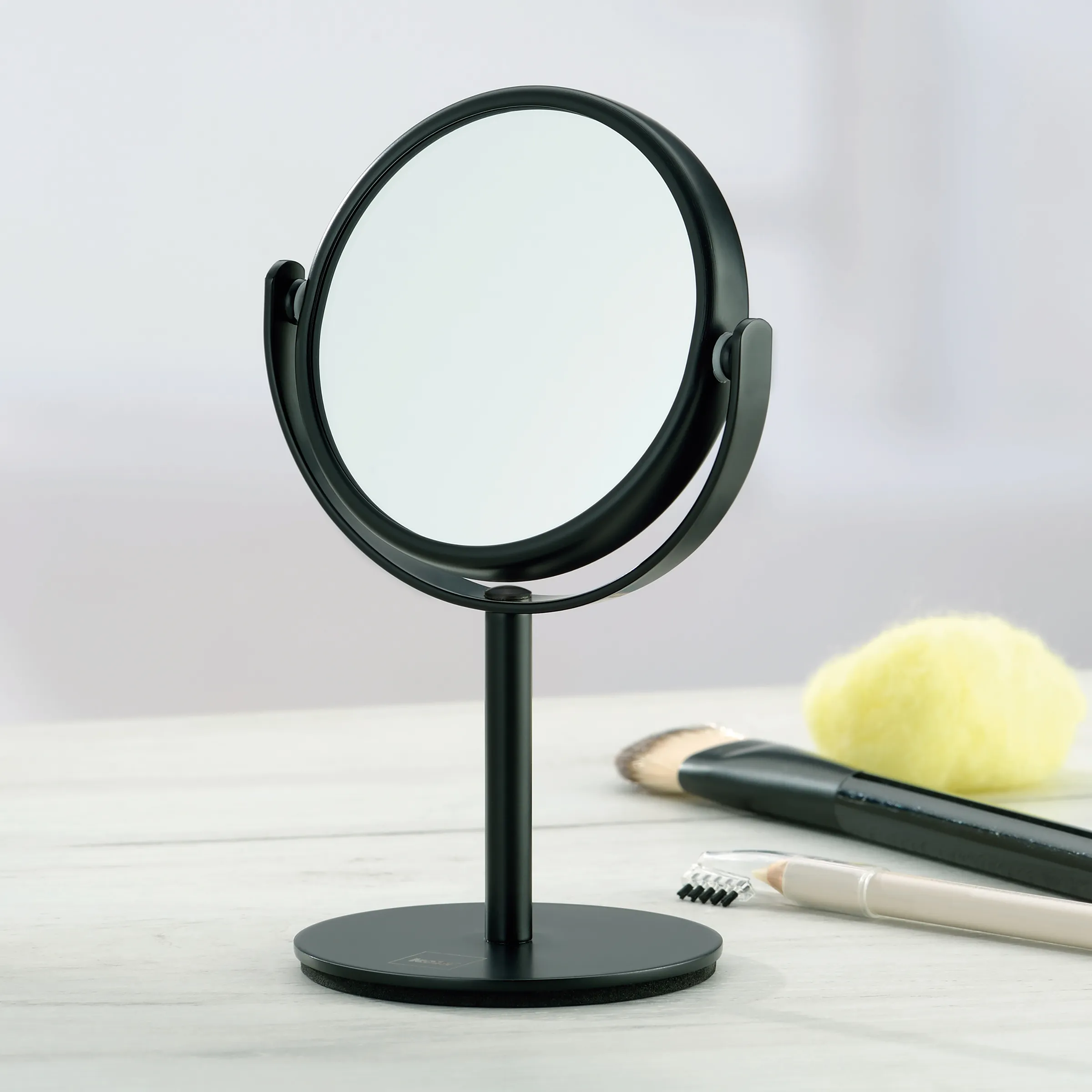 Standspiegel rund schwarz - Kosmetik Schminkspiegel mit Ständer Schwarzer Standspiegel mit rundem Rahmen und Metallständer auf weißem Tisch, daneben Kosmetikprodukte wie Pinsel und Applikator