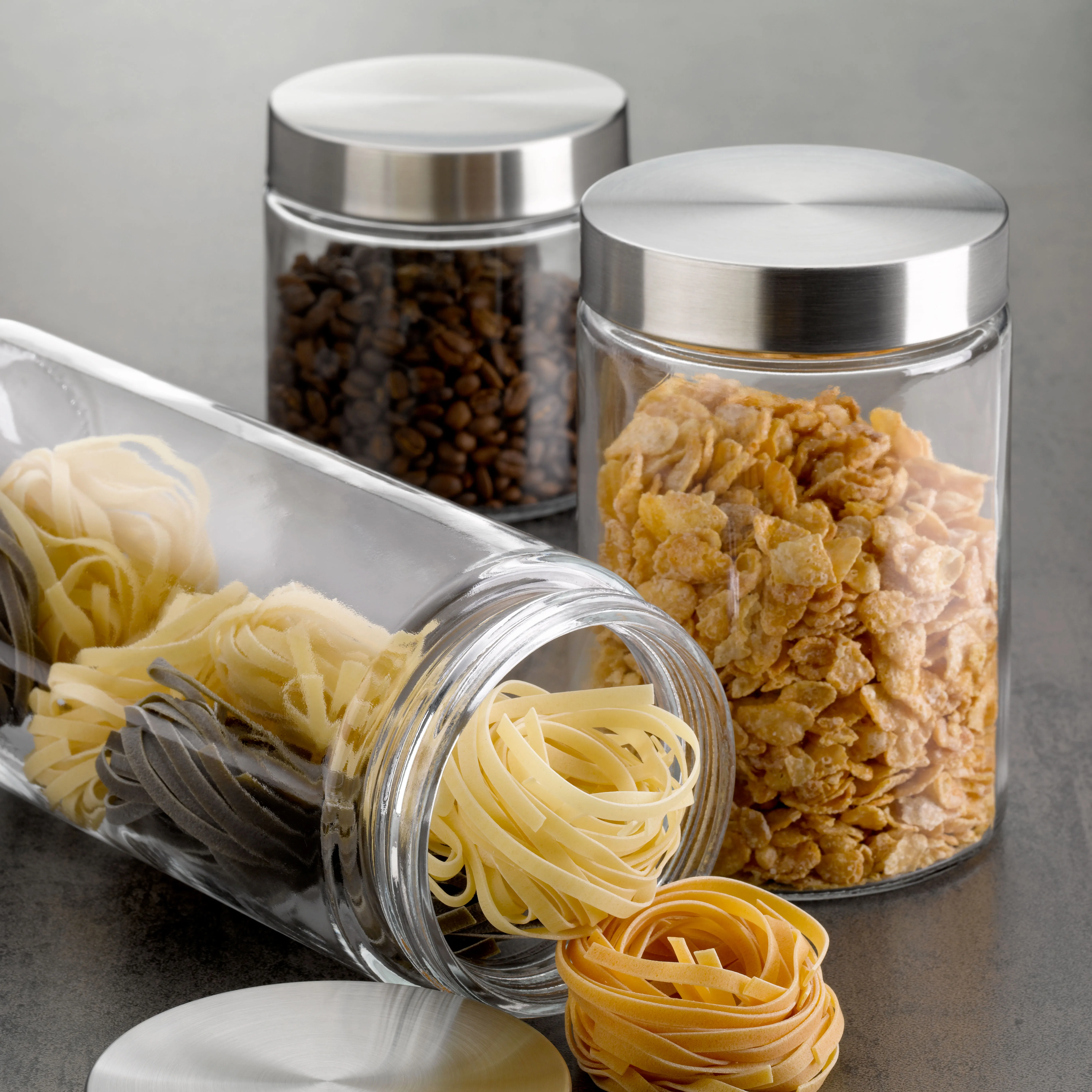 Vorratsgläser mit Edelstahldeckel gefüllt mit Nudeln, Cornflakes und Kaffeebohnen auf grauer Arbeitsfläche