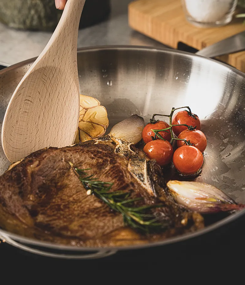 Holzlöffel rührt seared Steak in Pfanne mit Tomaten, Pilzen und Rosmarin während des Kochens