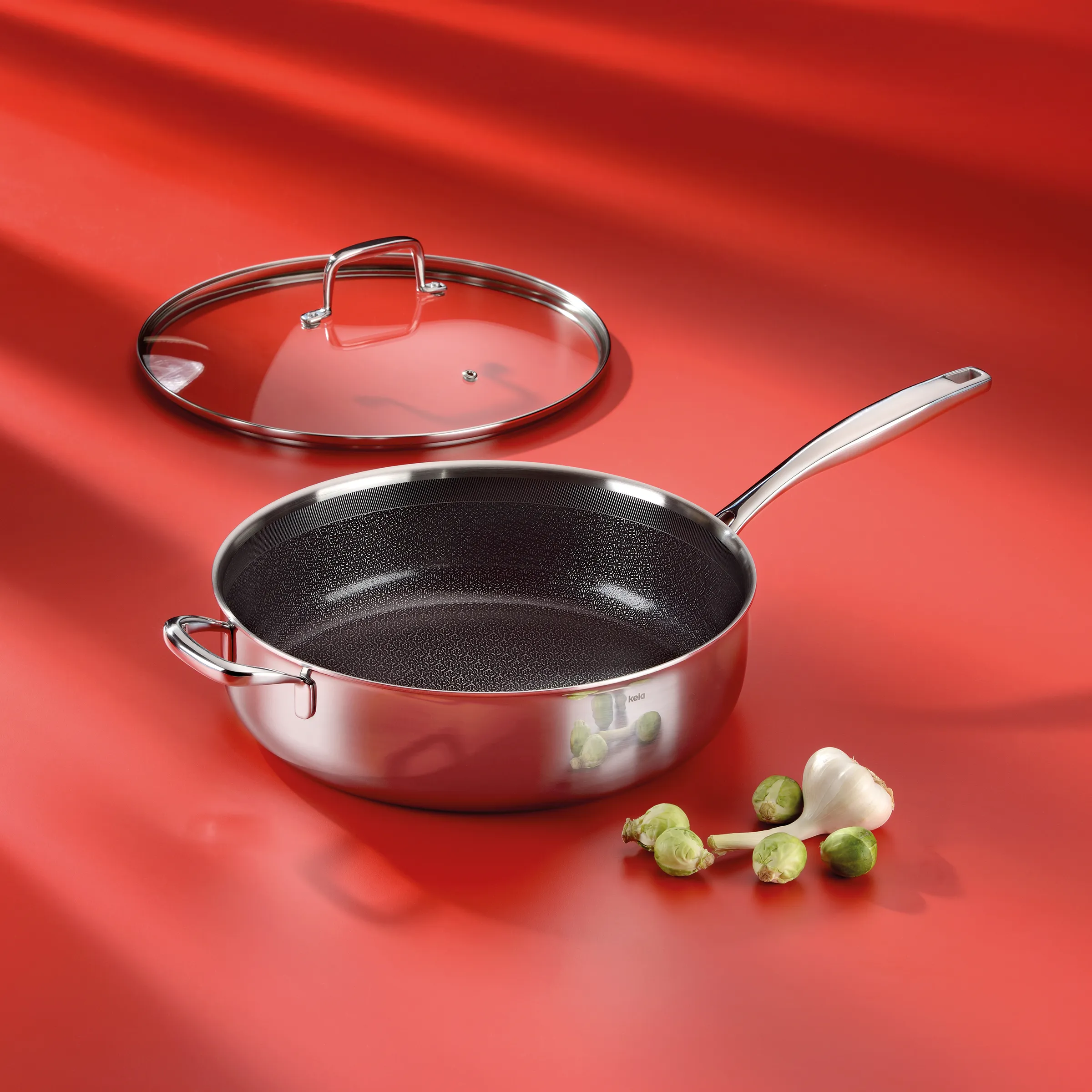 Sauteuse mit Glasdeckel und Antihaftbeschichtung - Edelstahl-Pfanne Edelstahl-Sauteuse mit Antihaftbeschichtung und Glasdeckel auf rotem Hintergrund mit frischem Knoblauch