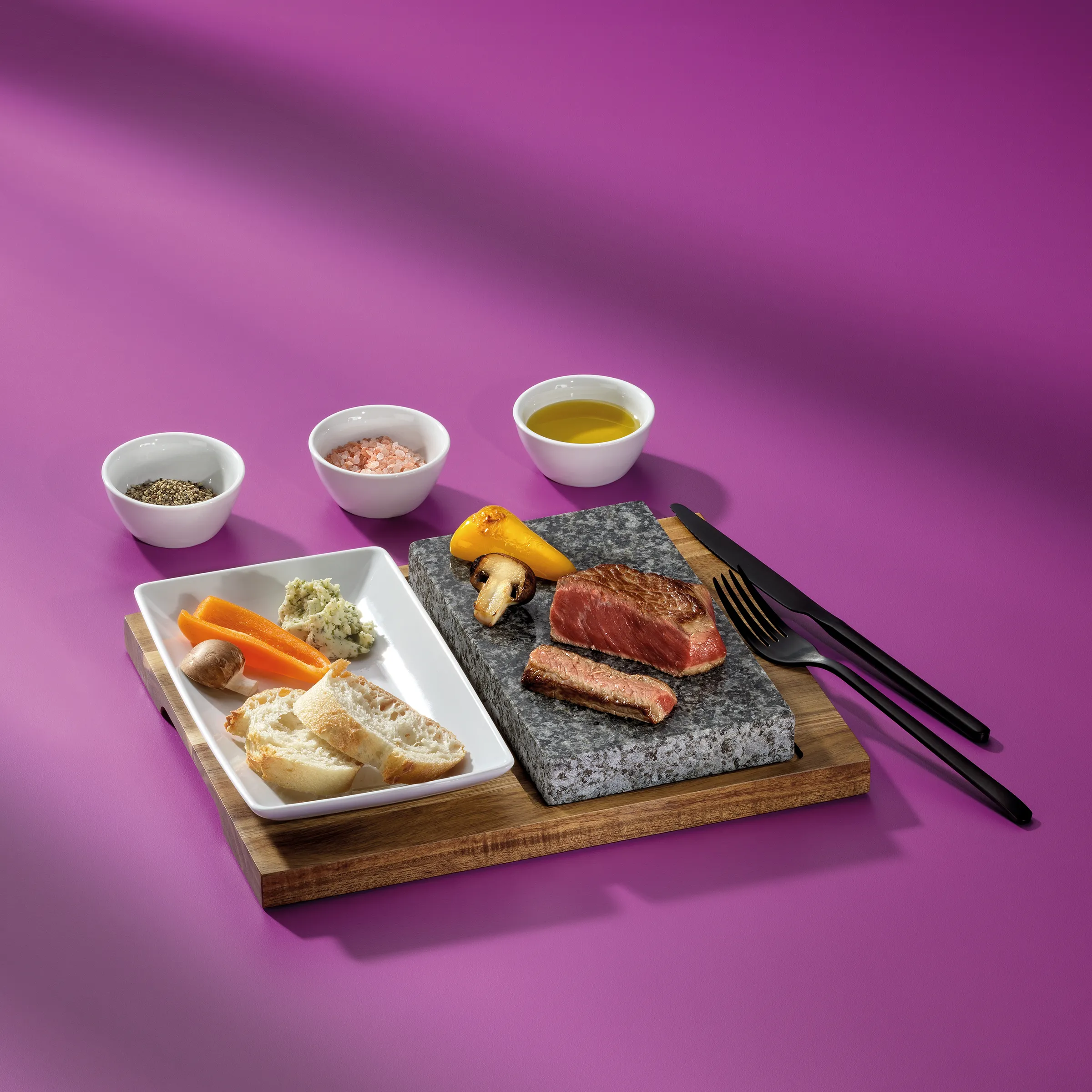 Gegrilltes Steak auf Steinplatte mit Gemüsebeilage und Gewürzen - Premium Food Fotografie Gegrilltes Steak auf Steinplatte mit Gemüse, Baguette, Olivenöl und Gewürzen auf Holzbrett vor rosa Hintergrund