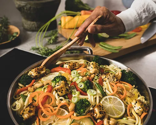 Asiatische Nudelpfanne - Rezept mit Gemüse und Wok Hände rühren eine Pfanne mit Asiatic Nudelauflauf, Gemüse und Limette um auf Schneidebrett mit Zutaten