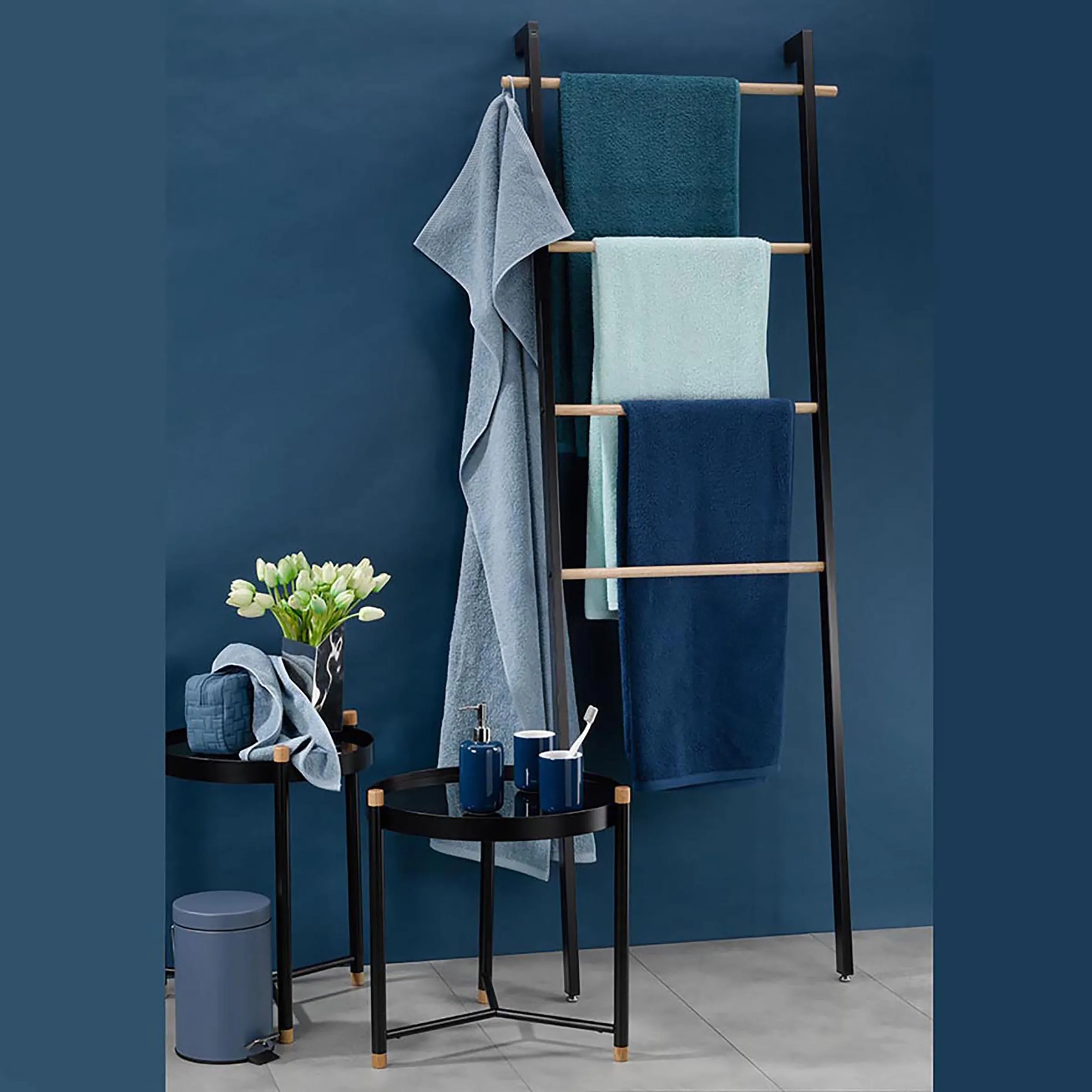 Handtuchständer mit Badaccessoires Set - Moderne Badezimmereinrichtung Modernes Badezimmer-Set mit schwarzem Handtuchständer, verschiedenfarbigen Handtüchern, schwarzem Beistelltisch mit Blumenvase und Badaccessoires vor blauer Wand