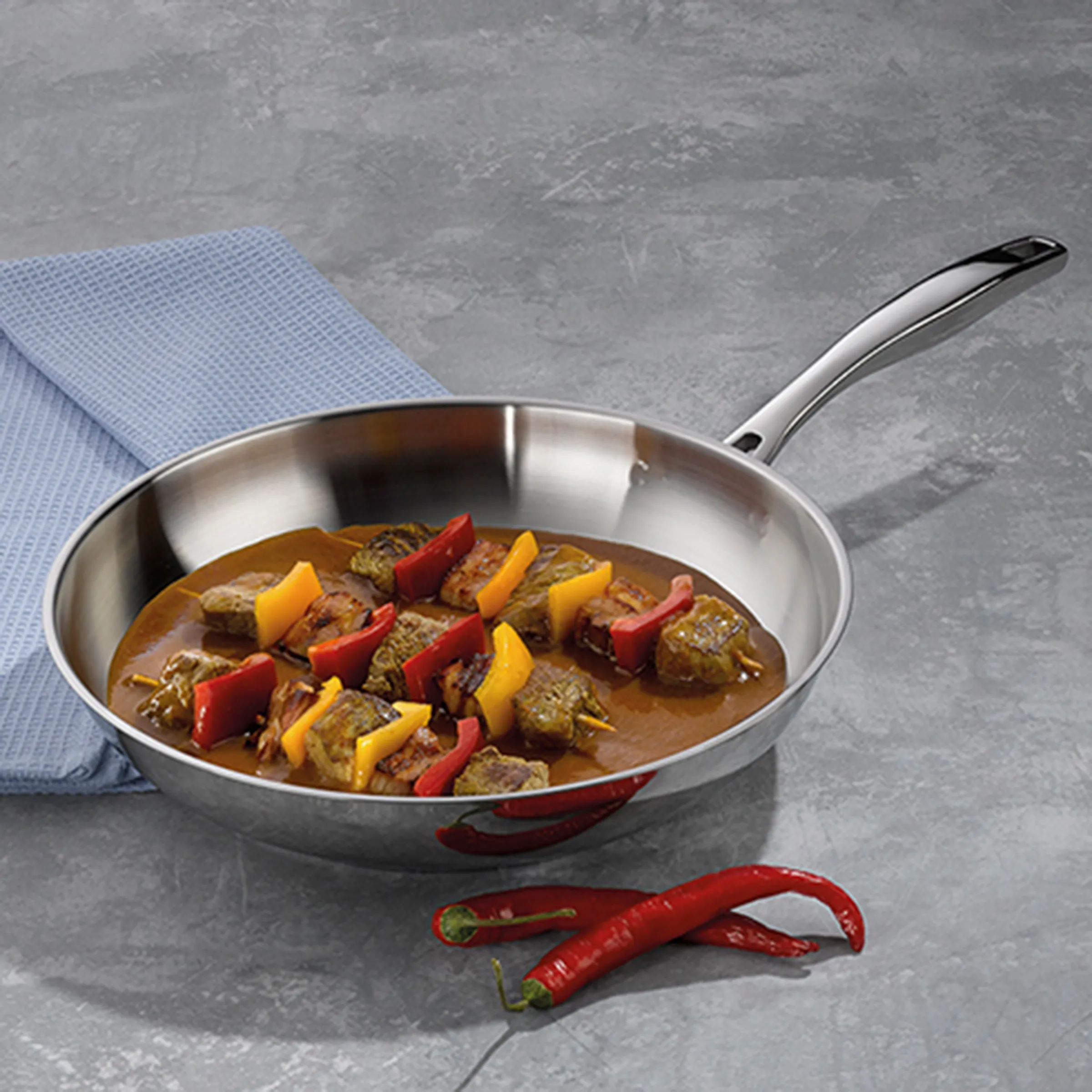 Bratpfanne mit Gulasch und Paprika - Rezept Kochgeschirr Edelstahl-Bratpfanne mit Fleischstücken, roten und gelben Paprika-Streifen in brauner Sauce auf grauem Untergrund