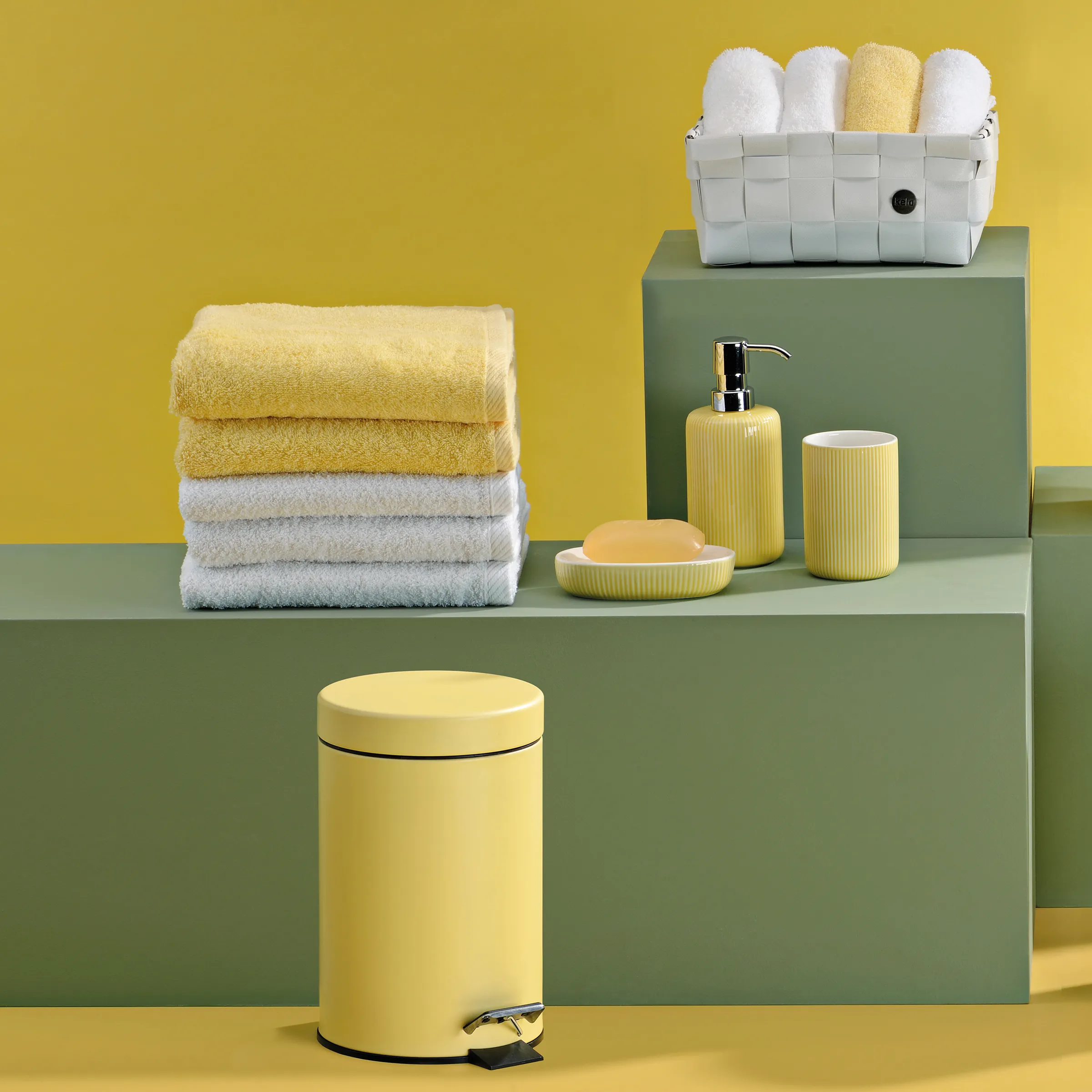 Badezimmer-Accessoires Set mit gelben und weißen Handtüchern, Seifenspender, Zahnbürstenbecher und gelbem Tretmülleimer auf grünem Podest.