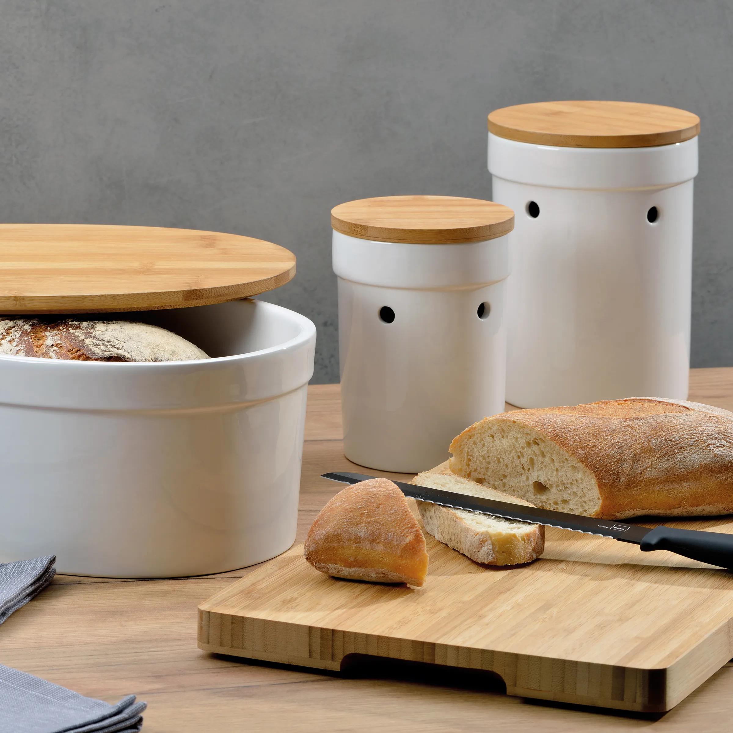 Weiße Brotdosen mit Bambusdeckel und Lüftungslöchern neben frischem Brot auf Holzbrett