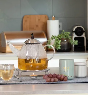 Glasteekannappe mit Tee auf grauem Tablett neben Trauben, Tasse und Kerze