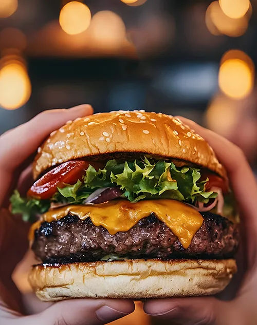 Premium Gourmet Burger mit Käse und Speck Hände halten einen saftigen Gourmet-Burger mit Sesambrötchen, Käse, Fleischpatty, Speck und grünem Salat vor unscharfem Hintergrund.