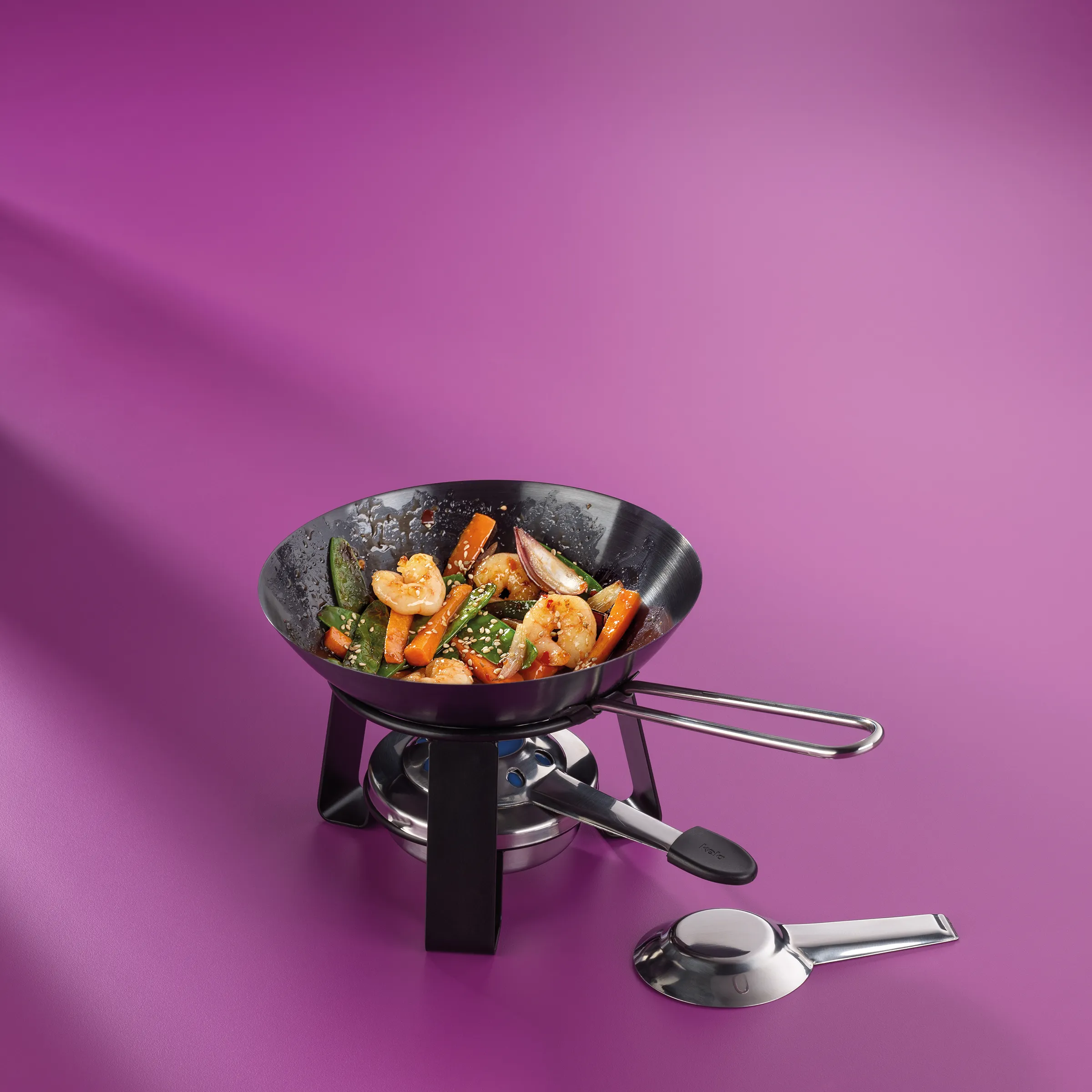Tragbarer Wok auf Gaskocher mit Shrimps, Gemüse und Soße auf violettem Hintergrund, daneben Servierlöffel
