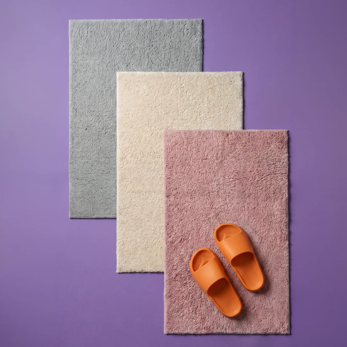 Drei textile Badematte in Grau, Beige und Mauve mit orangenen Hausschuhen auf violettem Hintergrund
