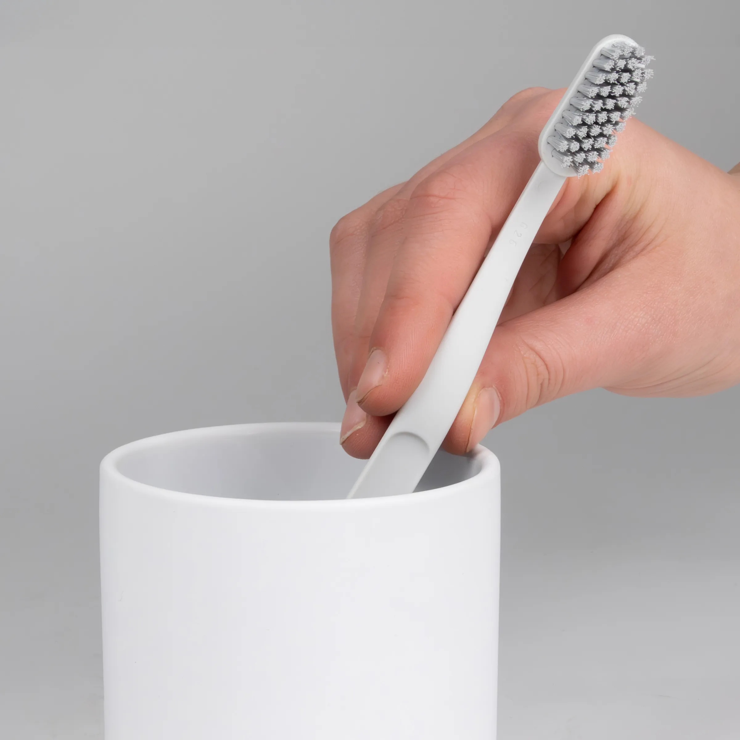 Hand hält weiße Zahnbürste mit Aktivkohlborsten über minimalistischem weißem Zahnbürstenhalter