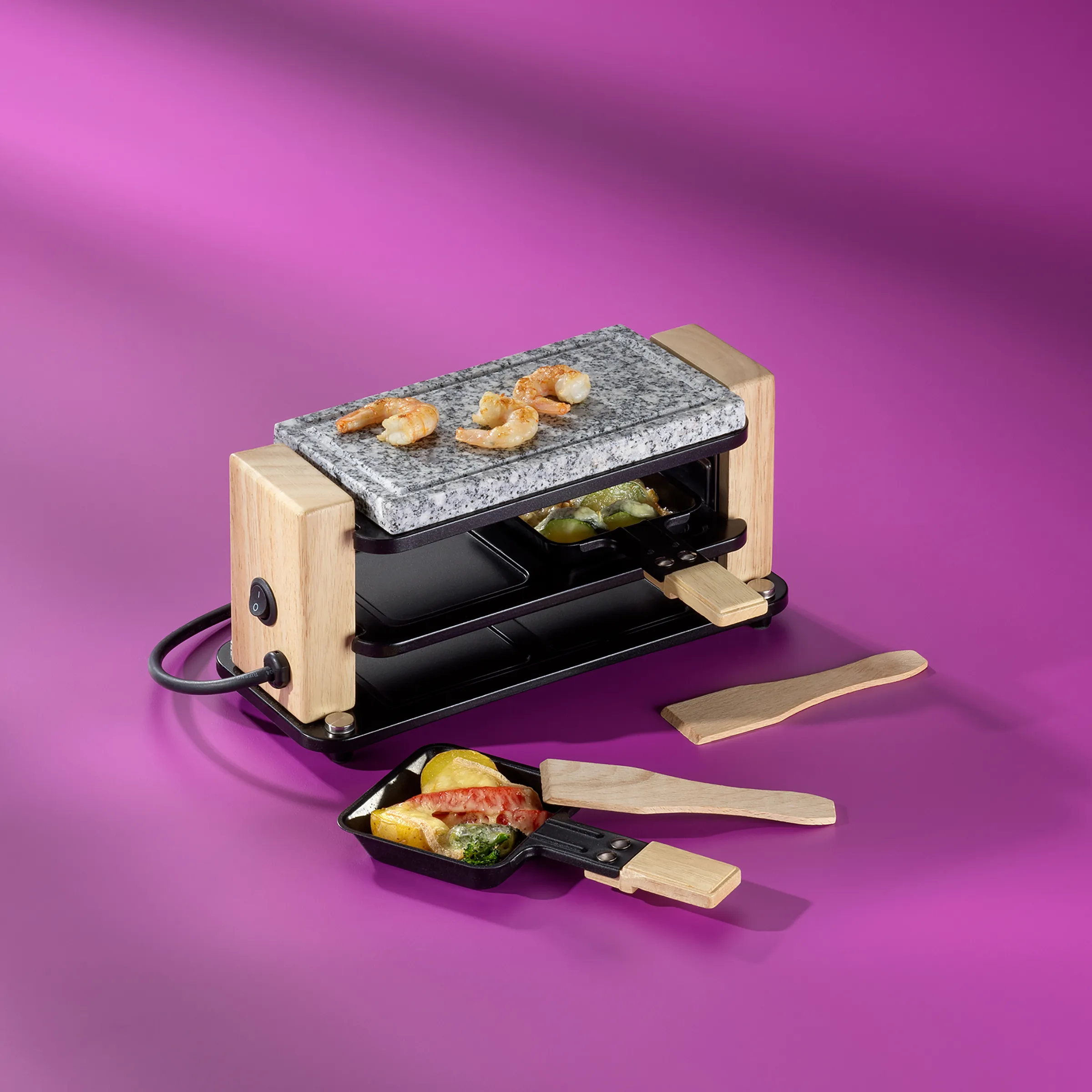 Raclette-Grill Set mit Granitplatte und Zubehör Raclette-Grill mit Holzrahmen, Granitplatte oben, Shrimps und Gemüse, schwarze Pfännchen mit Holzspachteln auf magentafarbenem Hintergrund