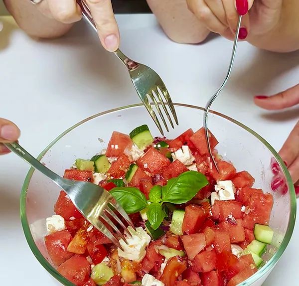 Drei Personen essen gemeinsam einen frischen Salat mit Wassermelone, Gurke, Feta und Basilikum aus einer Schüssel