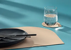 Modernes Tischgedeck mit Glasuntersetzer und Geschirr – Stilvolle Tischdekoration Glas Wasser auf beigem Untersetzer neben schwarzem Teller und Besteck auf türkisfarbenem Hintergrund