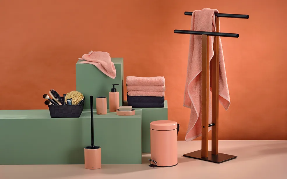 Badezimmer Accessoires Set - Handtuchständer und Badezimmerdeko Badezimmer-Set mit rosa Handtüchern, grünen Behältern, schwarzem Handtuchständer und Badaccessoires vor orangefarbenem Hintergrund