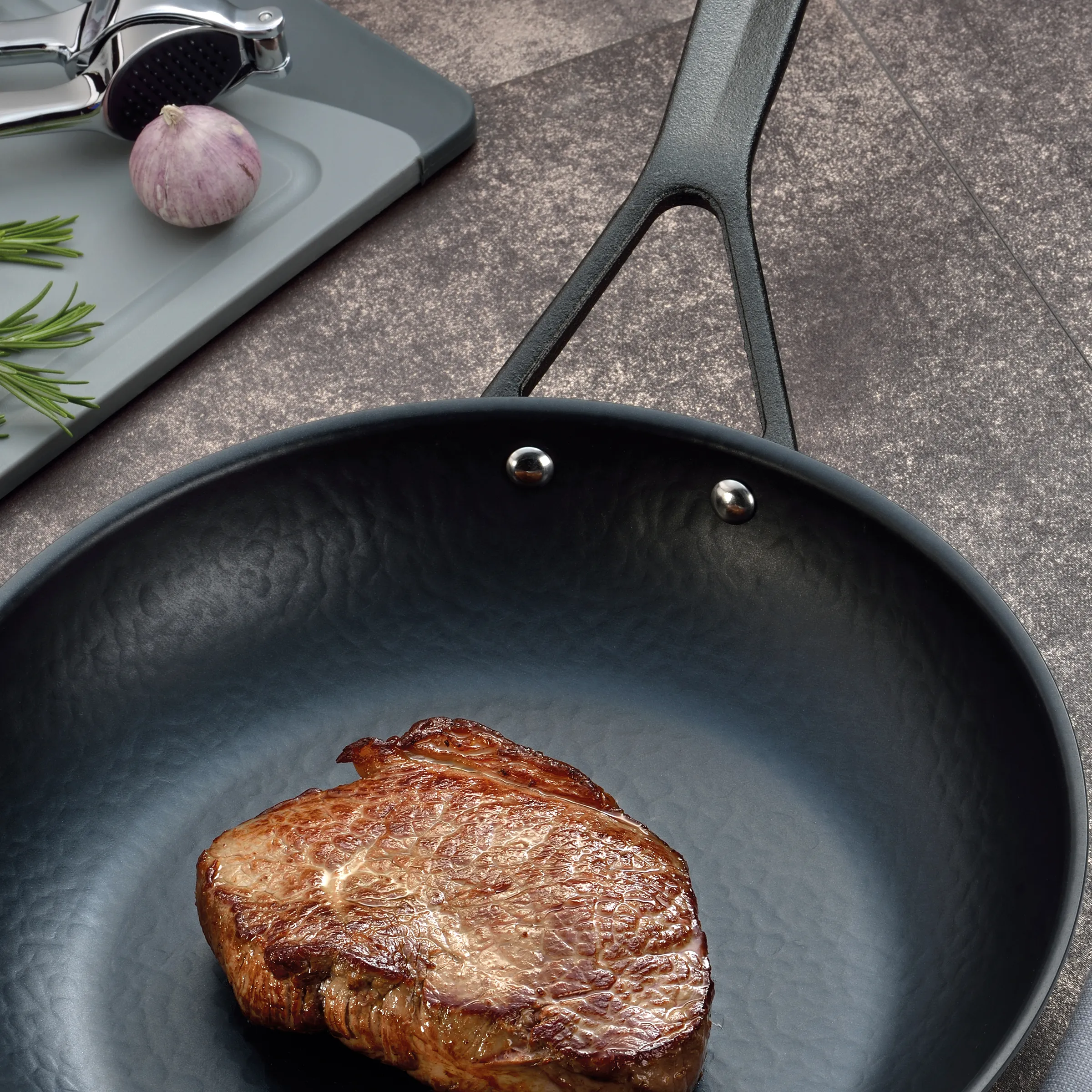 Steak braten: Premium-Fleisch in der Pfanne zubereiten Gebratenes Steak in schwarzer Pfanne mit Rosmarinzweigen und rosa Knoblauch auf grauem Untergrund