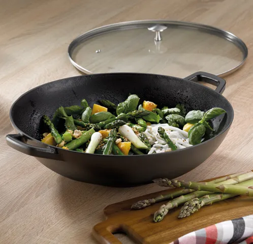 Wok-Pfanne mit Glasdeckel - Gemüsepfanne mit Spargel und frischem Grüngemüse Schwarze Wok-Pfanne mit Glasdeckel gefüllt mit gedünstetem grünem Gemüse und Spargel auf Holzbrett