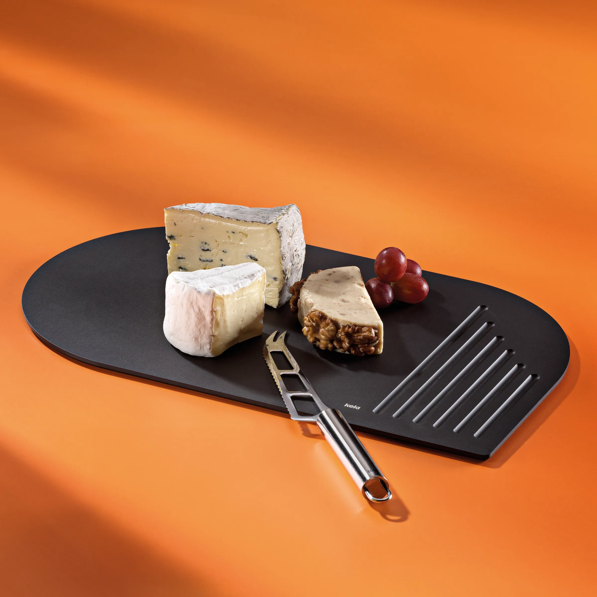 Käseplatte schwarz mit Käsemesser - Servierplatte für Käse und Snacks Schwarze ovale Käseplatte mit drei Käsesorten, Trauben und Käsemesser auf orangem Hintergrund