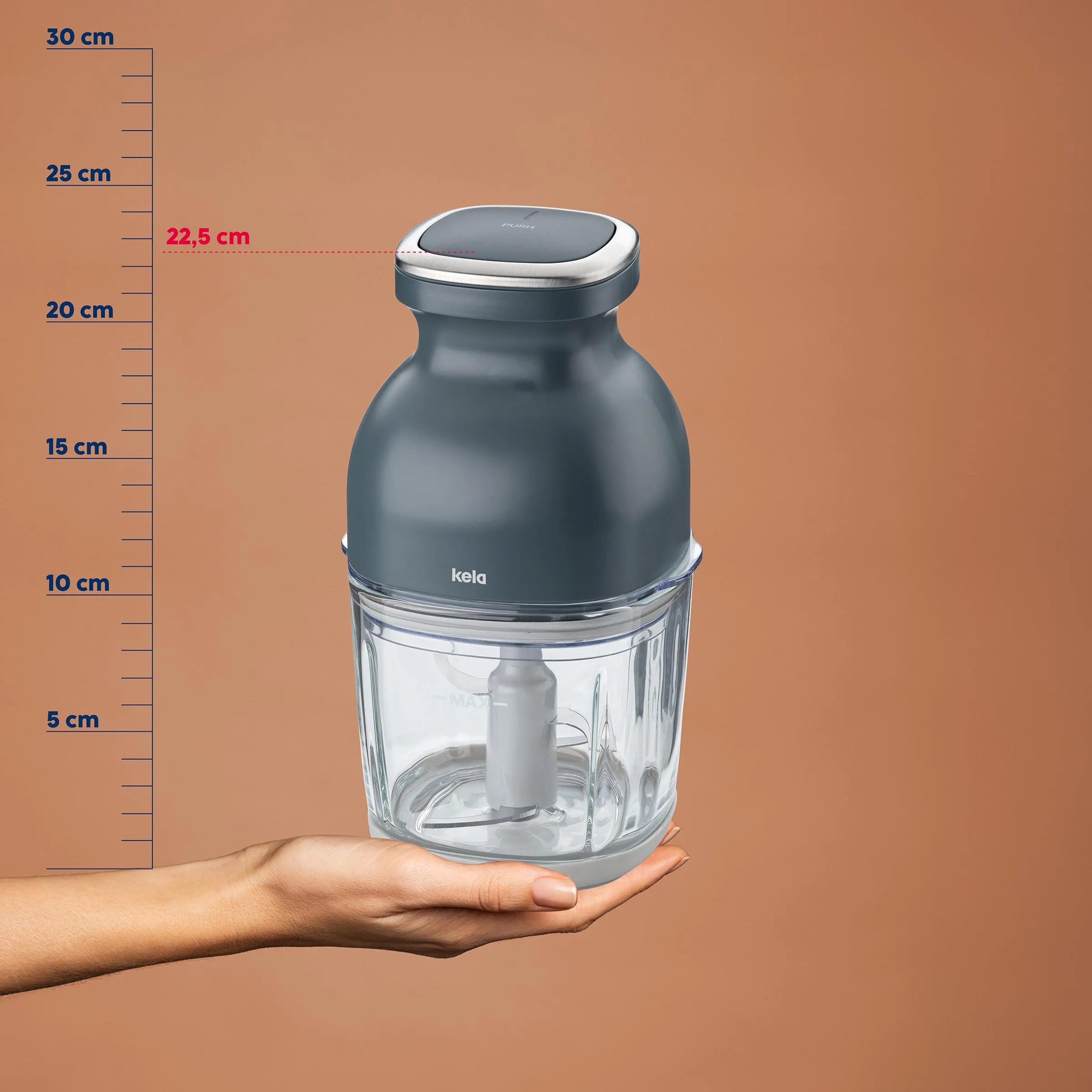 Kela Multizerkleinerer elektrisch mit Glasbehälter - kompakter Küchenhäcksler 22,5 cm Kela Zerkleinerer mit transparentem Behälter auf Hand, Größenskala im Hintergrund, 22,5 cm hoch