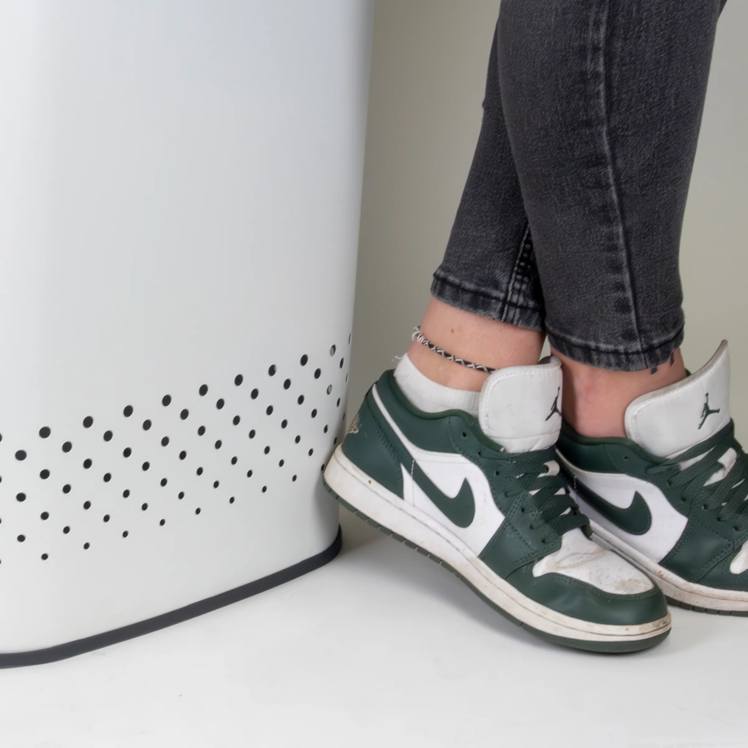 Person trägt dunkelgrüne und weiße Nike Air Jordan 1 Sneaker mit schwarzen Socken und schwarzer Jeans neben einem weißen Luftreiniger