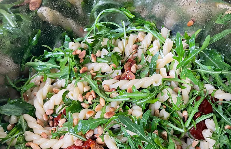 Pasta-Salat mit Rucola und Pinienkernen - Frischer Nudelsalat Spiralnudelsalat mit frischer Rucola, gerösteten Pinienkernen und getrockneten Tomaten auf weißem Teller