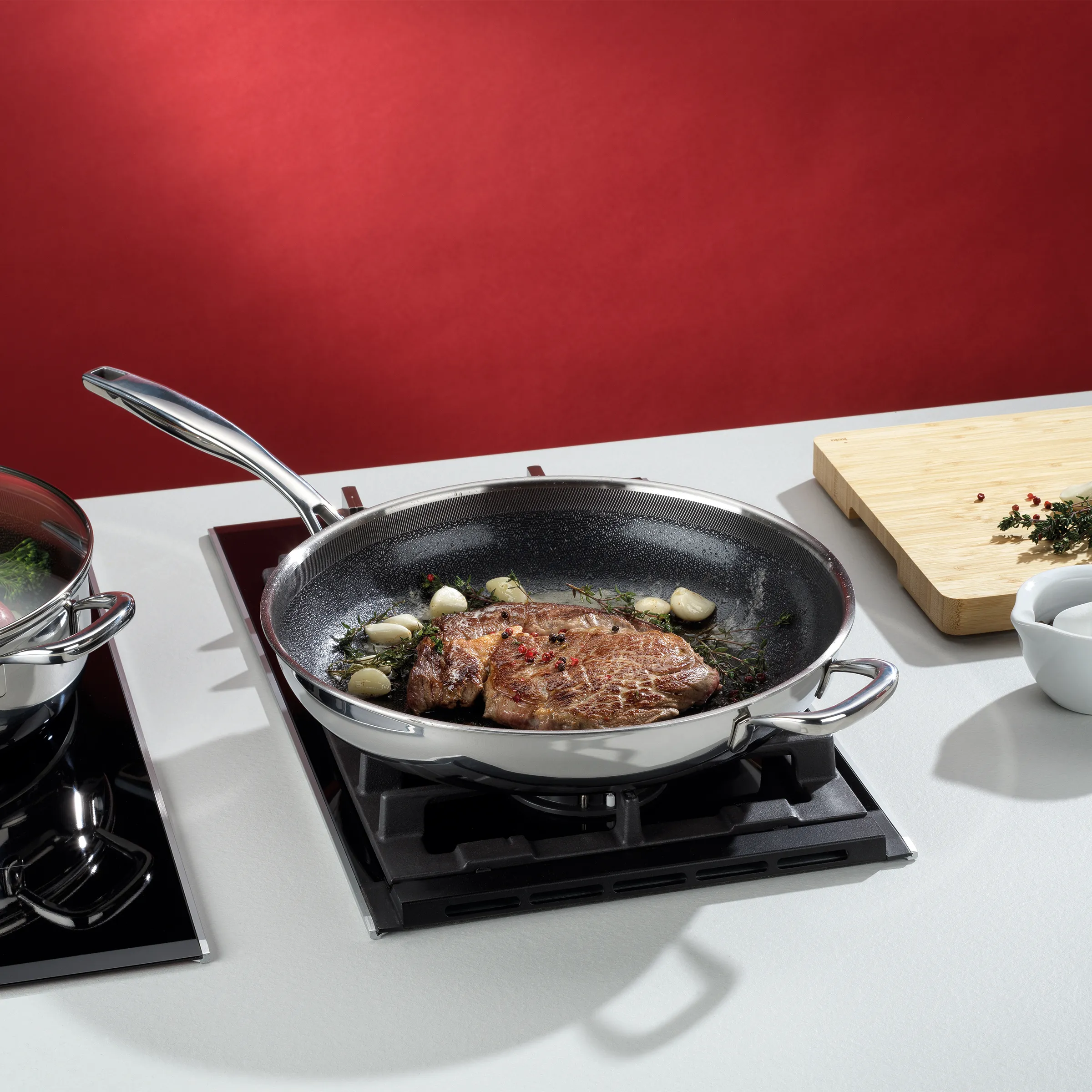 Braten braten in der Pfanne - Gourmet-Kochset Braten mit Pilzen und Kräutern in einer Pfanne auf schwarzem Kochfeld, neben Holzbrett und Kochutensilien