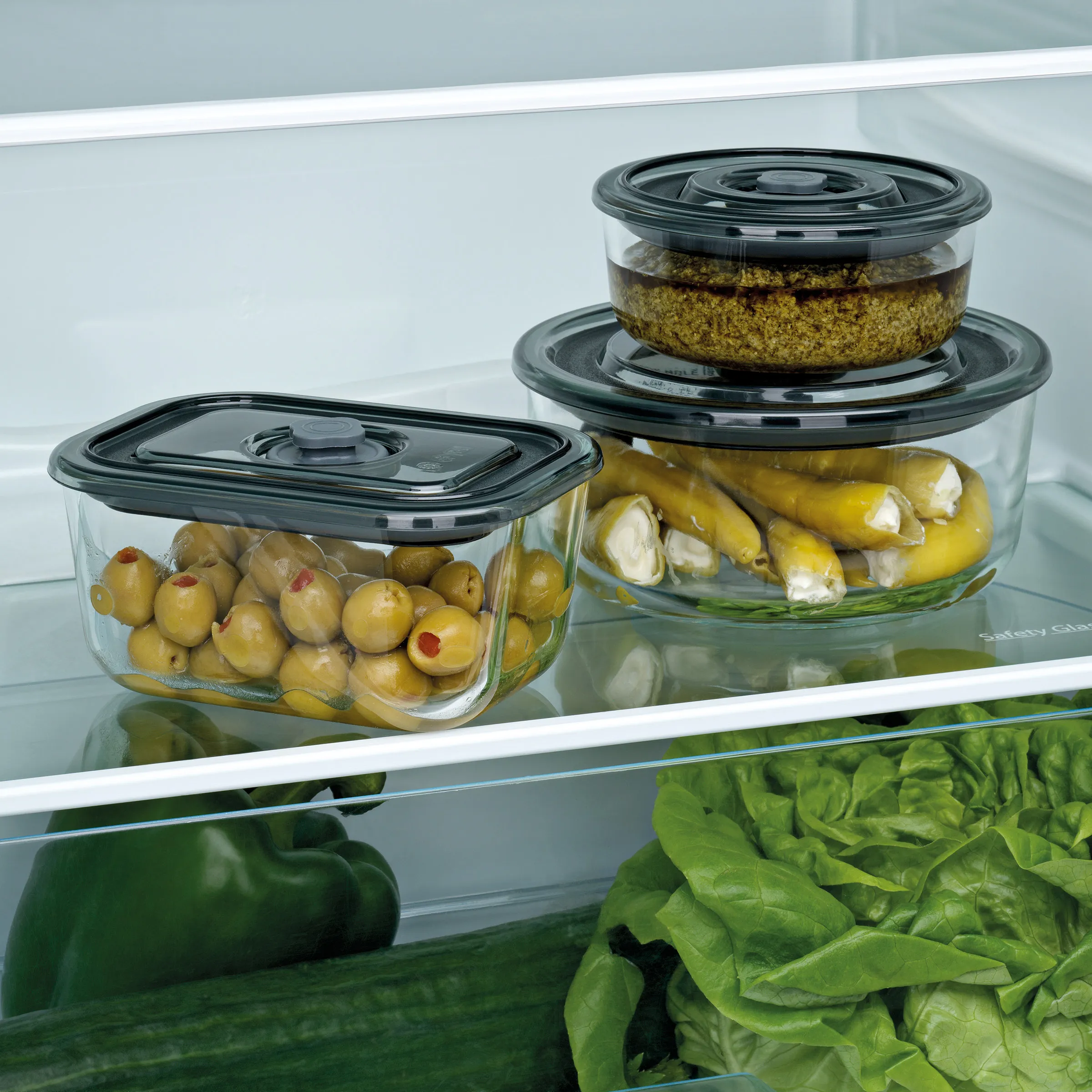 Glasbehälter mit Deckel für Lebensmittelaufbewahrung im Kühlschrank - frische Oliven und Gemüse Drei Glasbehälter mit Oliven, gefüllten Paprikaschoten und Pesto in einem Kühlschrank, daneben Salat