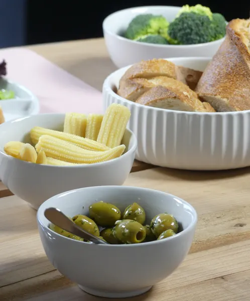 Servierschüsseln mit Gemüse und Brot - Tafelbesteck Weiße Schüsseln mit Brokkoli, Kartoffeln, Brot, grünen Oliven und anderen Lebensmitteln auf Holztisch.