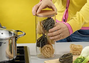 Hausgemachte Pasta aufbewahren - frische Nudeln im Glas mit Pastamaschine Person in gelber Jacke füllt frisch zubereitete schwarze und helle Nudeln in Glas mit Pastamaschine im Hintergrund