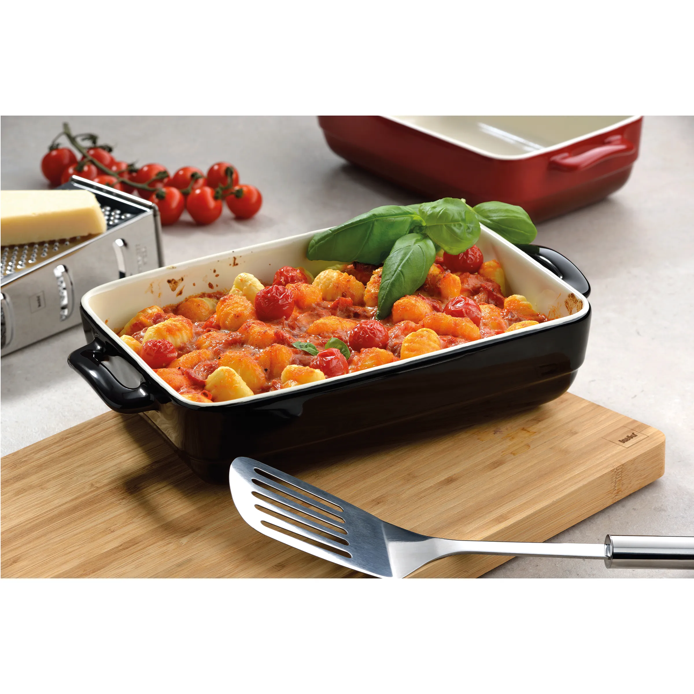 Gnocchi-Auflaufform mit Tomatensauce - Emaille-Backform für italienische Pasta Gnocchi mit Tomatensauce und Basilikum in schwarzer Auflaufform auf Holzbrett mit Käsereibe und Tomaten