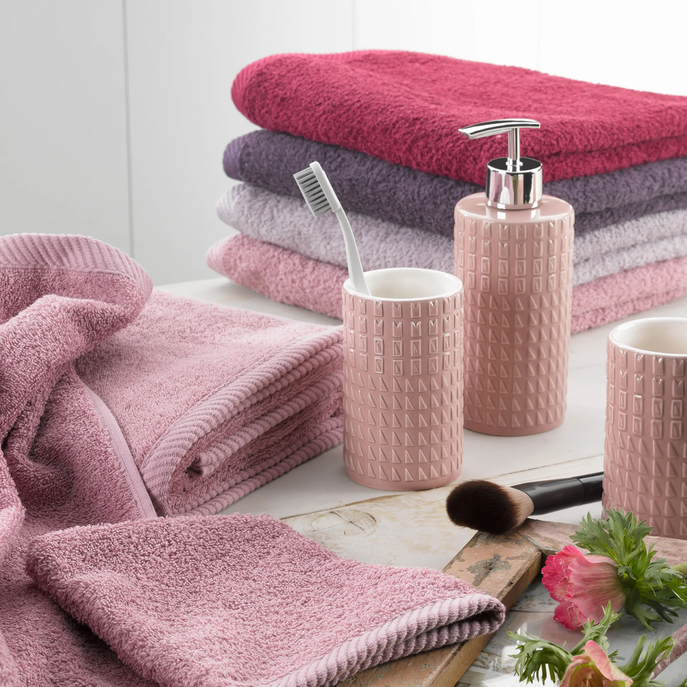 Badezimmer-Set mit rosa Keramikbehältern, Zahnbürste, Kosmetikpinsel und gestapelten Handtüchern in verschiedenen Farben