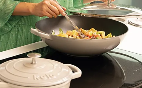 Person rührt buntes Gemüse in einem schwarzen Wok mit Holzspatel um, neben weißem Keramik-Deckel auf Kochfeld