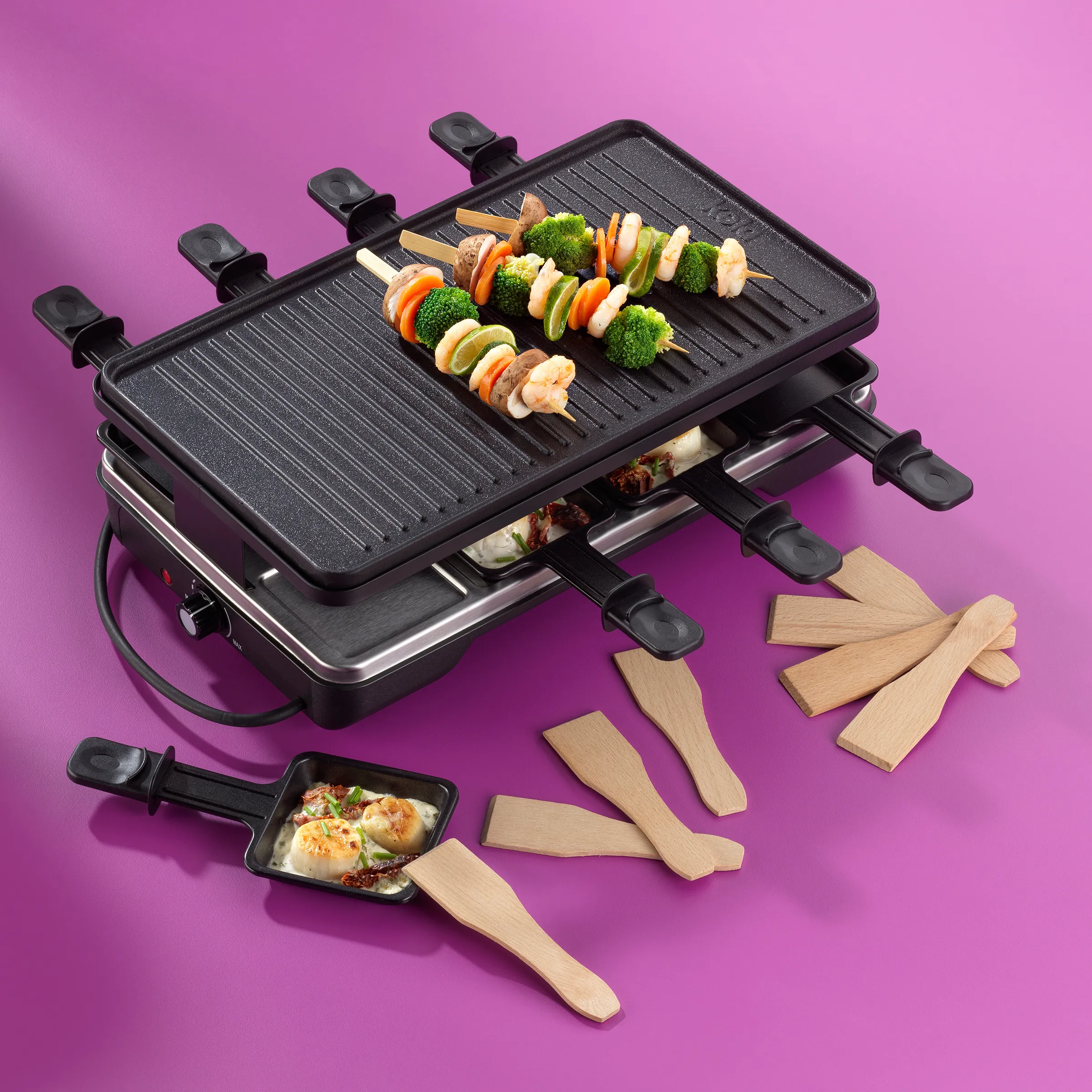 Raclette Grill Set mit Spachteln und Zubehör Raclette-Grill mit vier Holzspachteln, gefüllten Mini-Pfannen und grilliertem Gemüse-Spieß auf rosa Hintergrund
