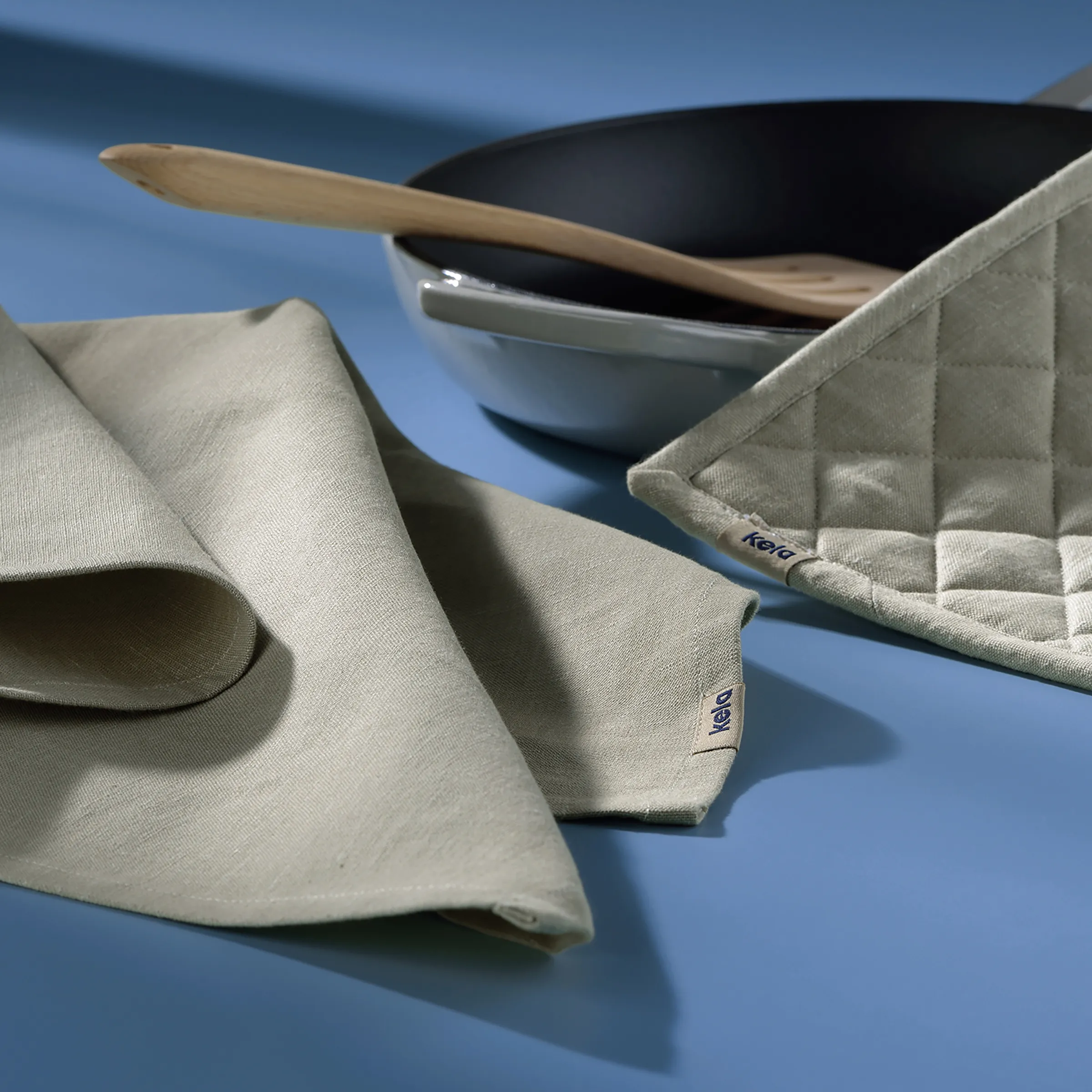 Hochwertige Leinen Geschirrtücher - Baumwolle & Design Beige Baumwoll-Geschirrtücher mit Label neben schwarzer Schale und Holzlöffel auf blauem Hintergrund