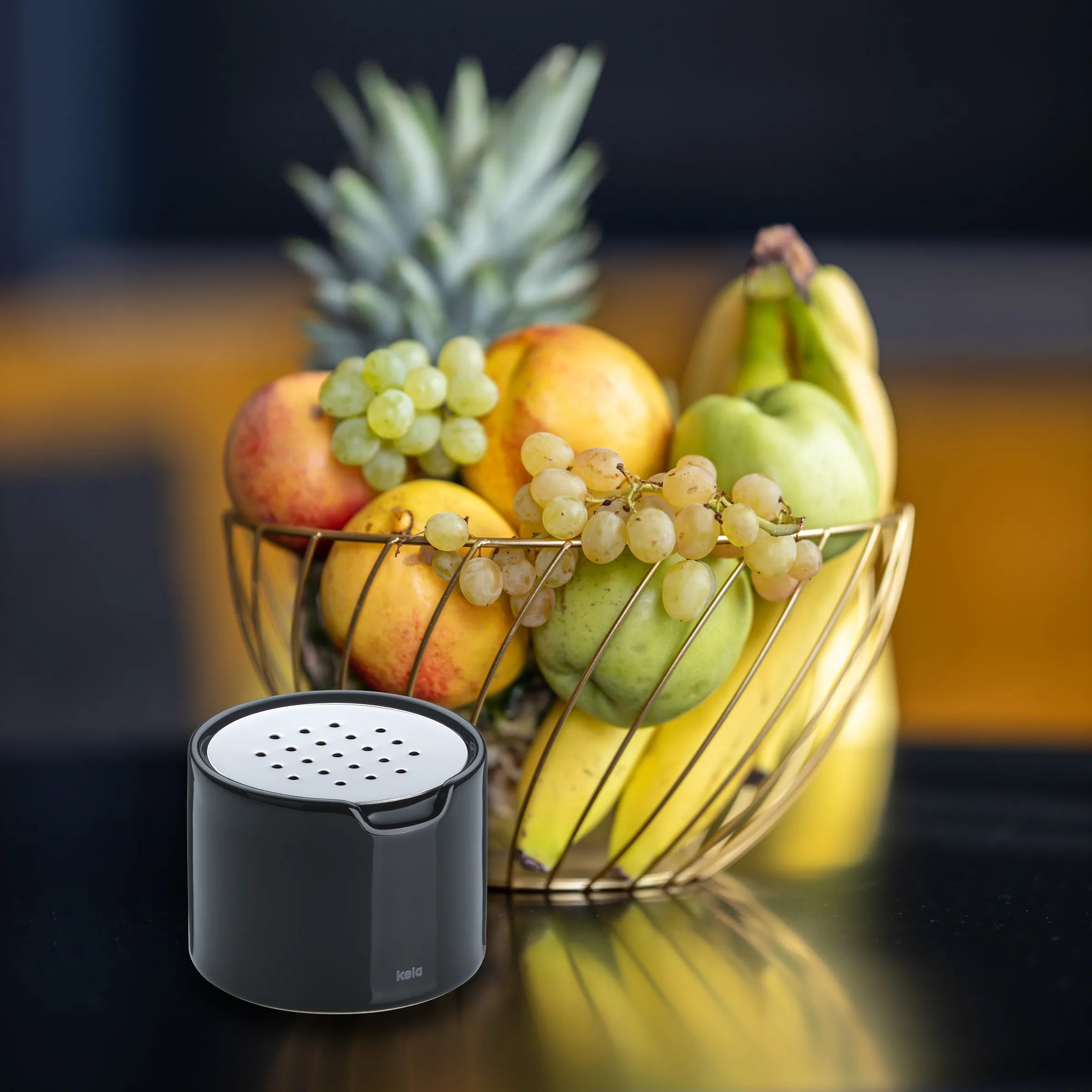 Kela Aktivkohlefilter für Obstkorb - Geruchsneutralisierung Küche Schwarzer Kela Aktivkohlefilter vor Obstkorb mit Bananen, Ananas, Trauben und Äpfeln auf Küchentheke