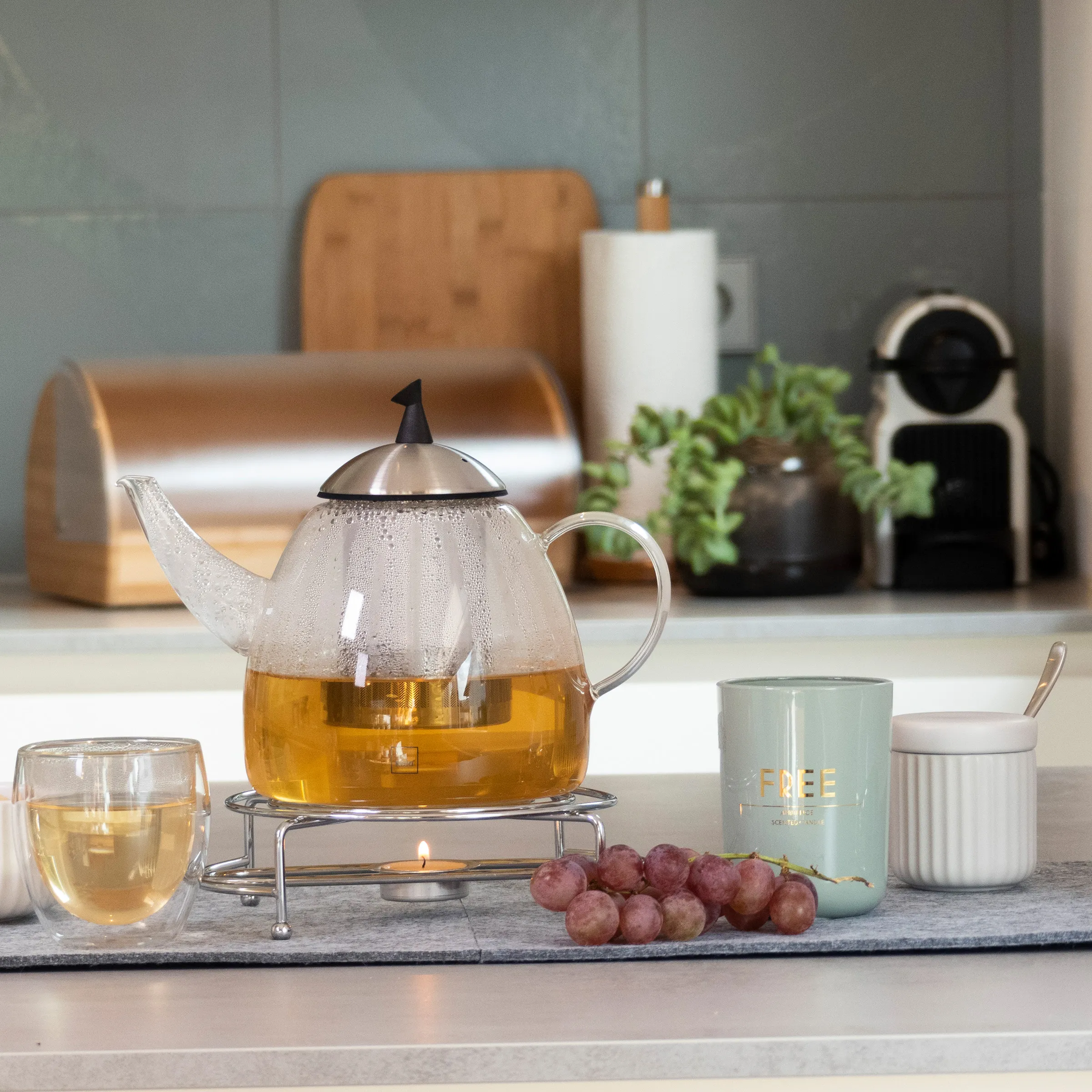 Glas-Teekanne mit Stövchen und Tee-Zubehör in moderner Küche Glas-Teekanne mit goldenem Tee auf Stövchen, daneben Teetasse, Weintrauben und Teedose auf Marmorplatte