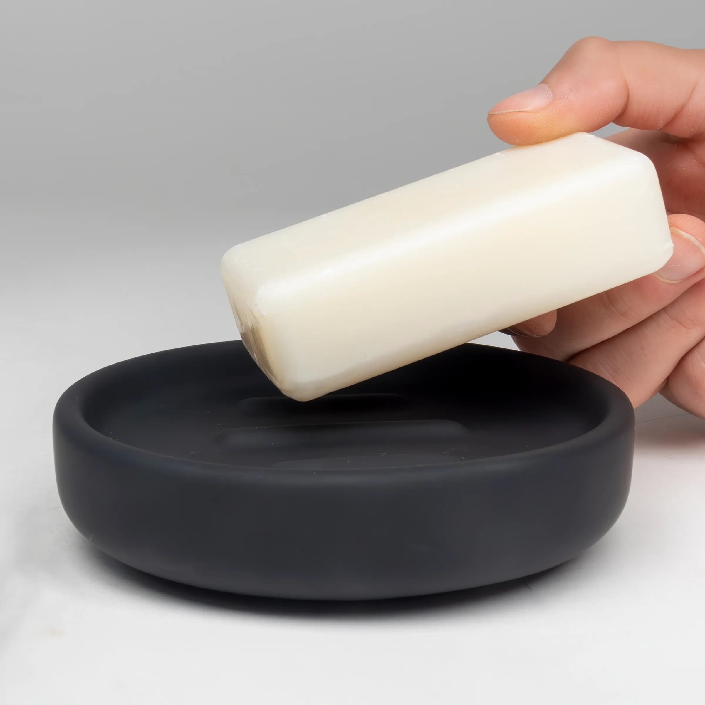 Weiße Seife und schwarze Seifenschale - Badezimmer Accessoires Hand hält eine weiße Seife über einer schwarzen Seifenschale vor grauem Hintergrund