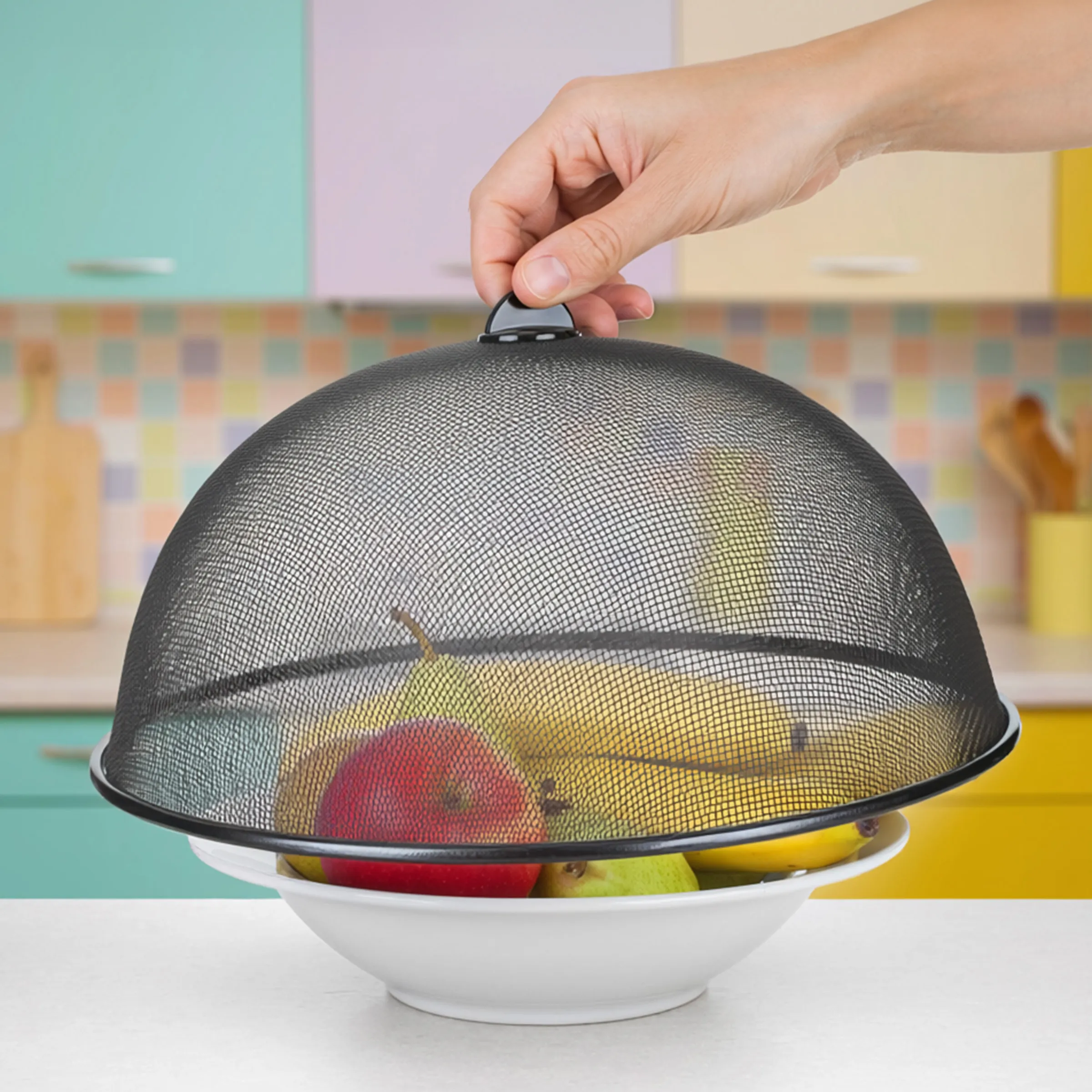 Obst Schutzhaube - Speisehaube für Küche Hand hebt schwarze Schutzhaube von einer weißen Schale mit frischem Obst in farbenfroher Küche