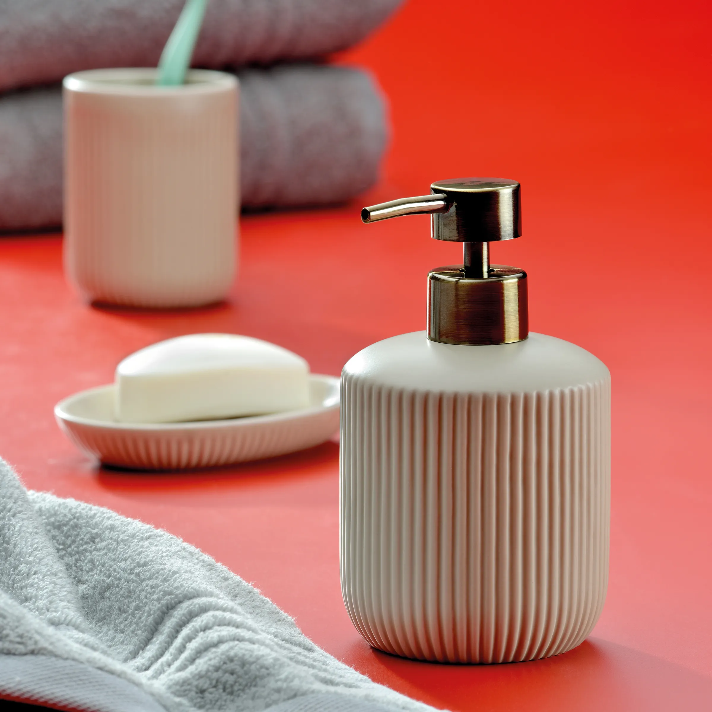 Badezimmer Accessoires Set beige - Seifenspender und Seifenschale Badezimmer-Set mit beigefarbenem Seifenspender, Seifenschale, Zahnputzbecher und grauen Handtüchern auf rotem Hintergrund
