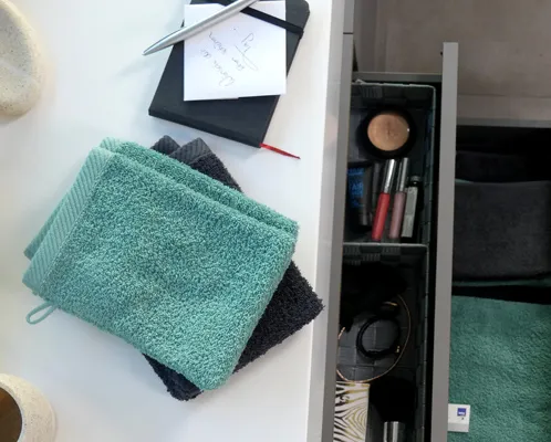 Türkise und schwarze Handtücher auf weißem Untergrund neben offener Schublade mit Kosmetik und Accessoires