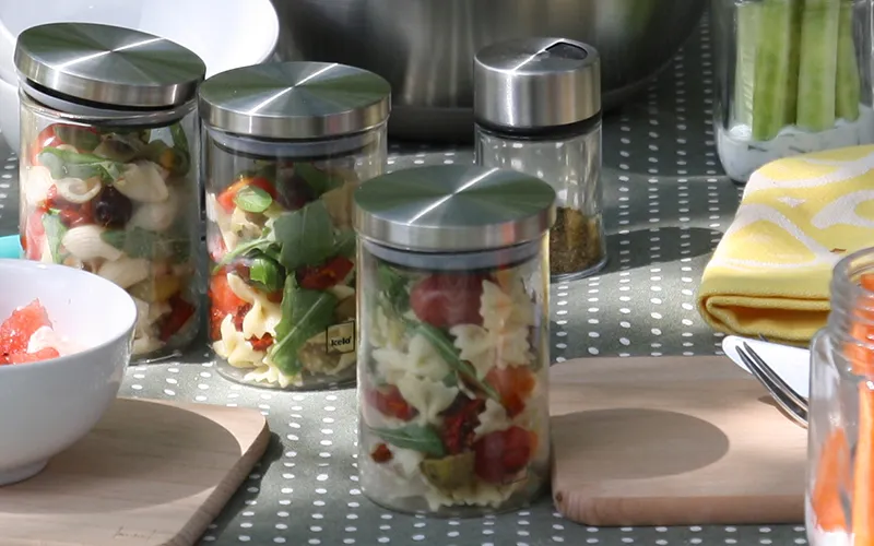 Meal Prep Salatboxen - Frische Salate in Glasbehältern Vier Glasbehälter mit Salatmischungen aus Gemüse, Kräutern und Dressing auf einem gepunkteten Tablett neben einer weißen Schüssel