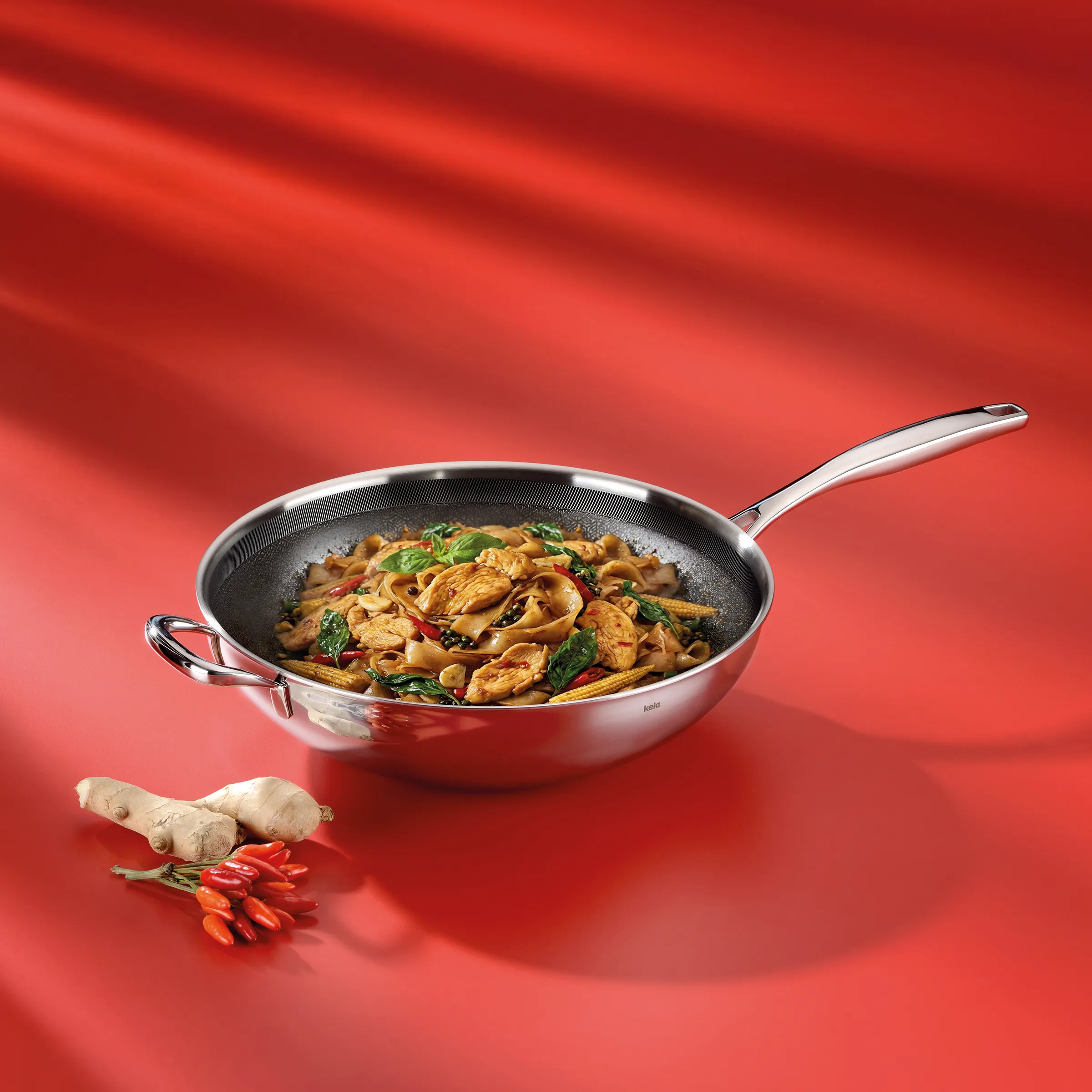 Wok Pfanne mit asiatischen Nudeln und Gemüse Edelstahl-Wok mit Nudel-Gemüse-Pfanne auf rotem Hintergrund, daneben Ingwer und rote Chilischoten
