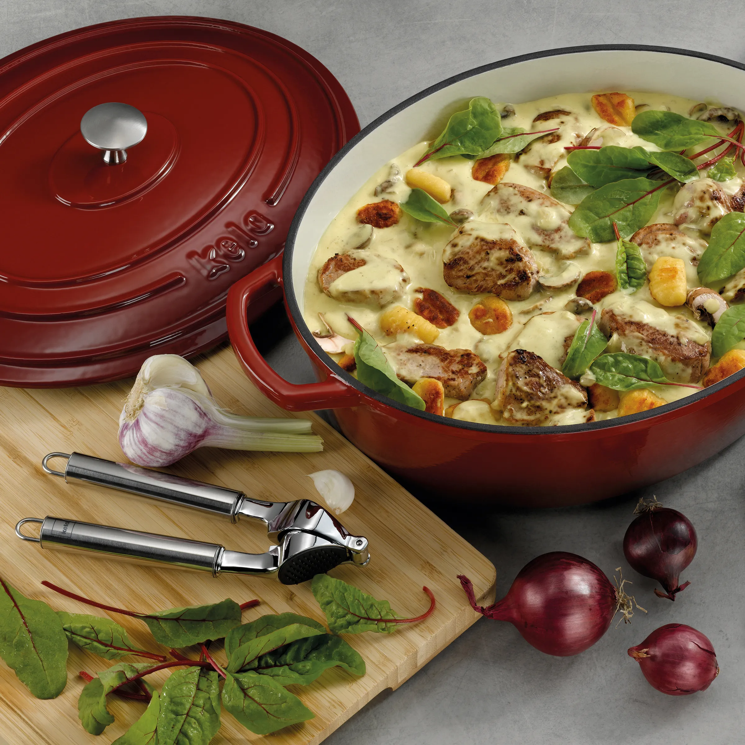 Roter Gusseisen-Bräter mit Deckel, gefüllt mit Gnocchi, Fleisch und Salat, daneben Knoblauch und Zwiebeln