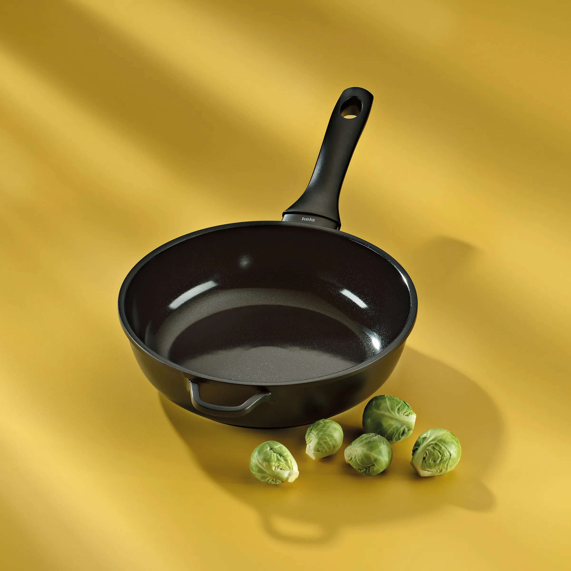 Beschichtete Bratpfanne mit Antihaftbeschichtung - Kela Kochgeschirr Schwarze Bratpfanne von Kela mit ergonomischem Griff auf gelbem Hintergrund, daneben fünf Rosenkohl