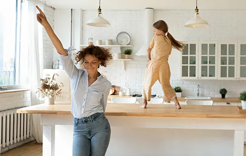 Glückliche Frauen tanzen in moderner Küche - Lebensfreude zuhause Zwei fröhliche Frauen tanzen ausgelassen in moderner weißer Küche mit Holzarbeitsplatte und hellen Hängelampen