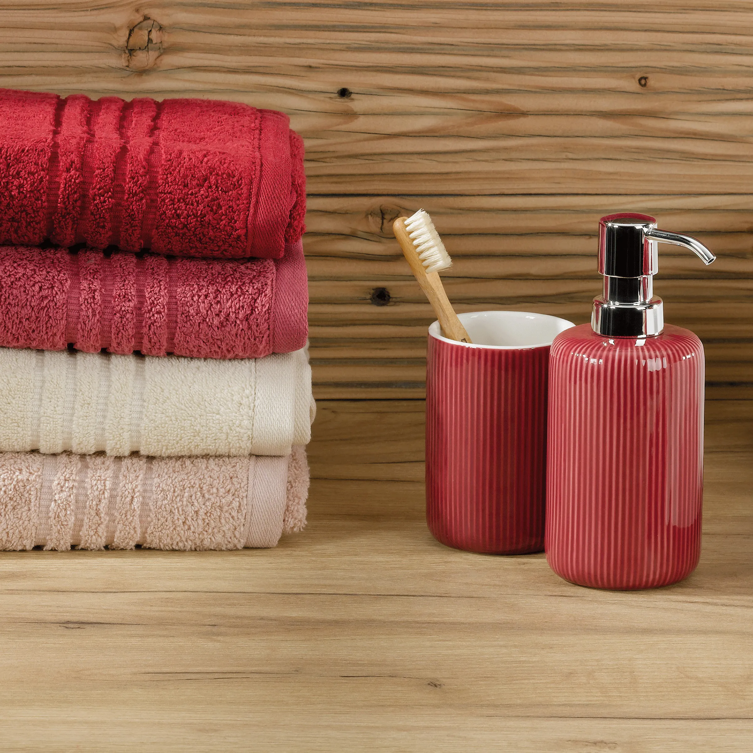 Badezimmer Accessoires Set rot Handtücher Badezimmer-Set mit roten und beigen Handtüchern, roten Behältern, Zahnbürste und Seifenspender auf Holzuntergrund.